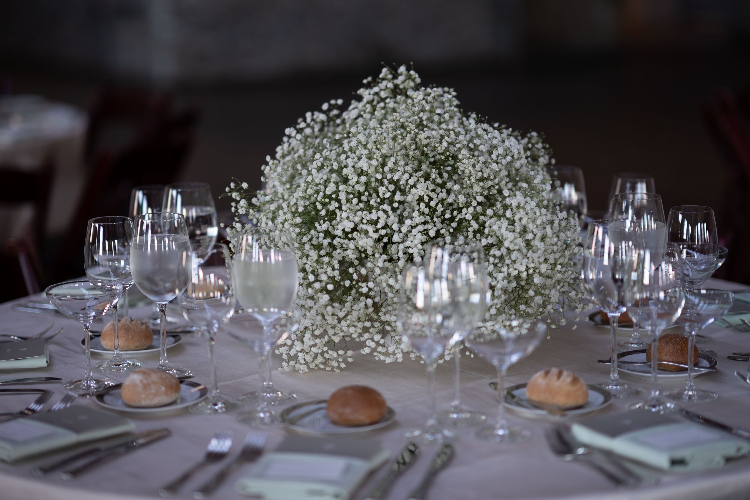 Elegant banquet table with a large centerpiece of white flowers, multiple wine glasses, bread rolls, and formal tableware.