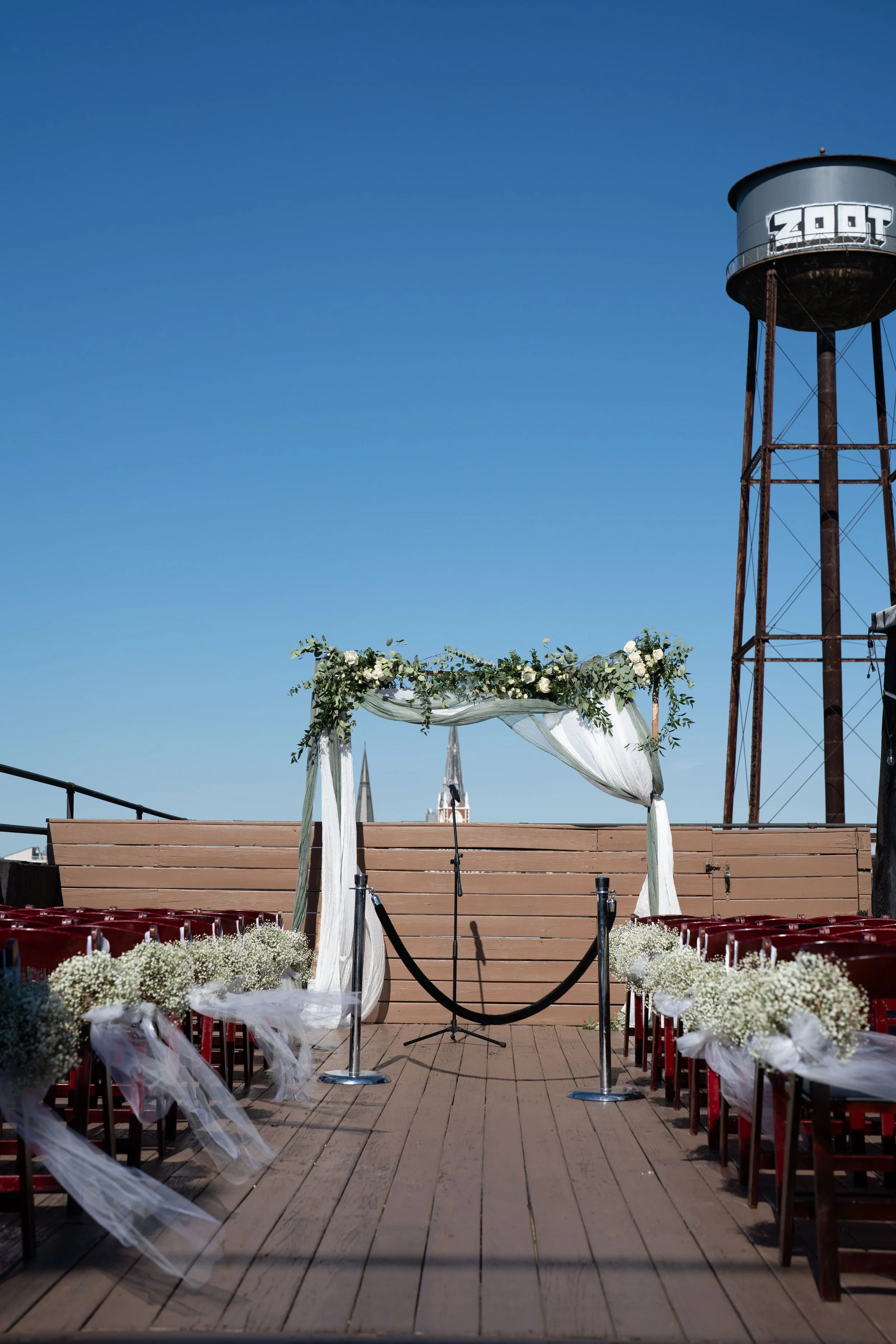 Rooftop wedding ceremony setup with an arch decorated with flowers and white drapery, red chairs with white floral arrangements on the sides, and a blue sky background, with a water tower labeled 'Zoot' in the distance.