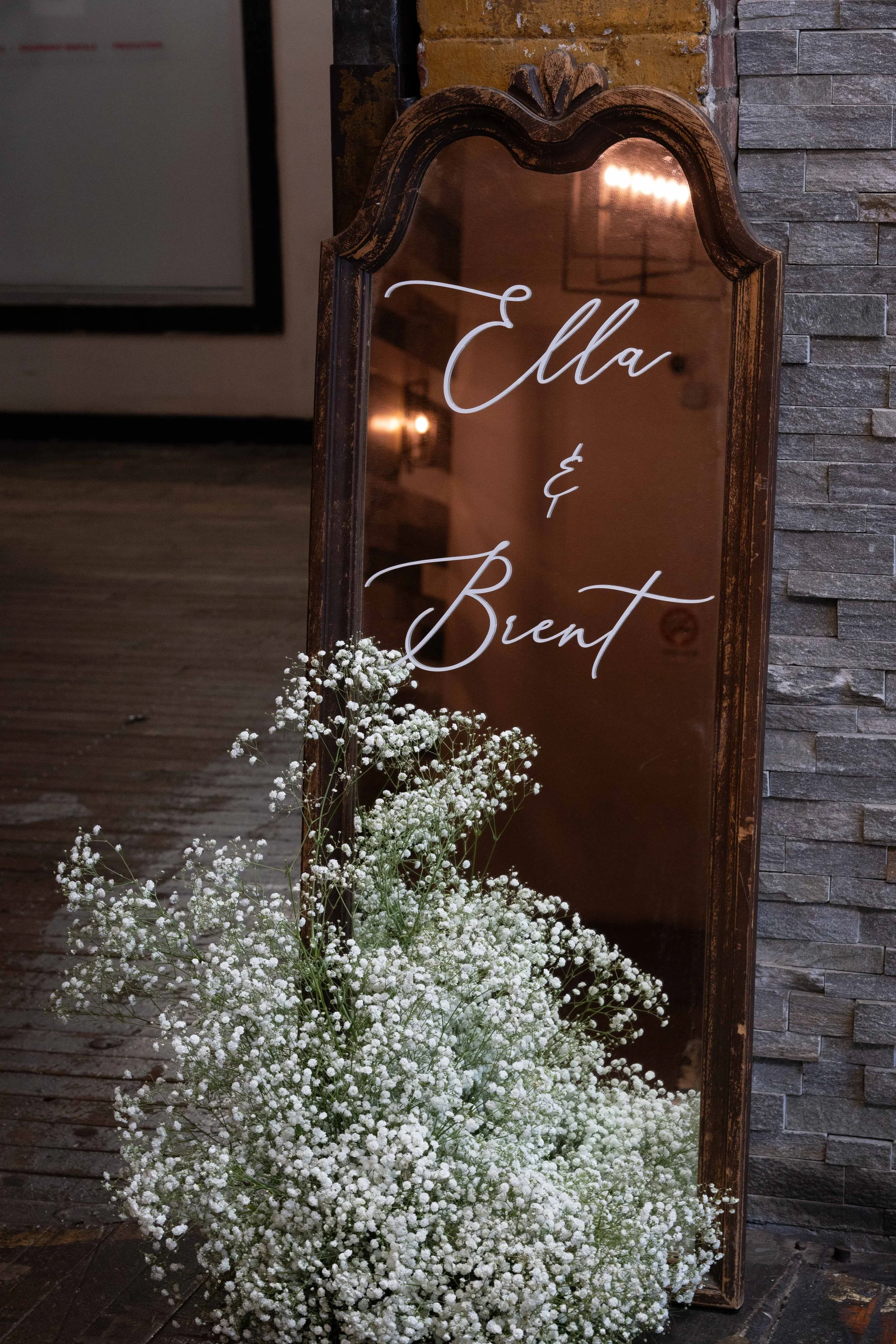 A vintage wooden framed mirror with the words 'Ella & Bient' written in cursive on it, decorated with white baby's breath flowers at the base, leaning against a brick wall.