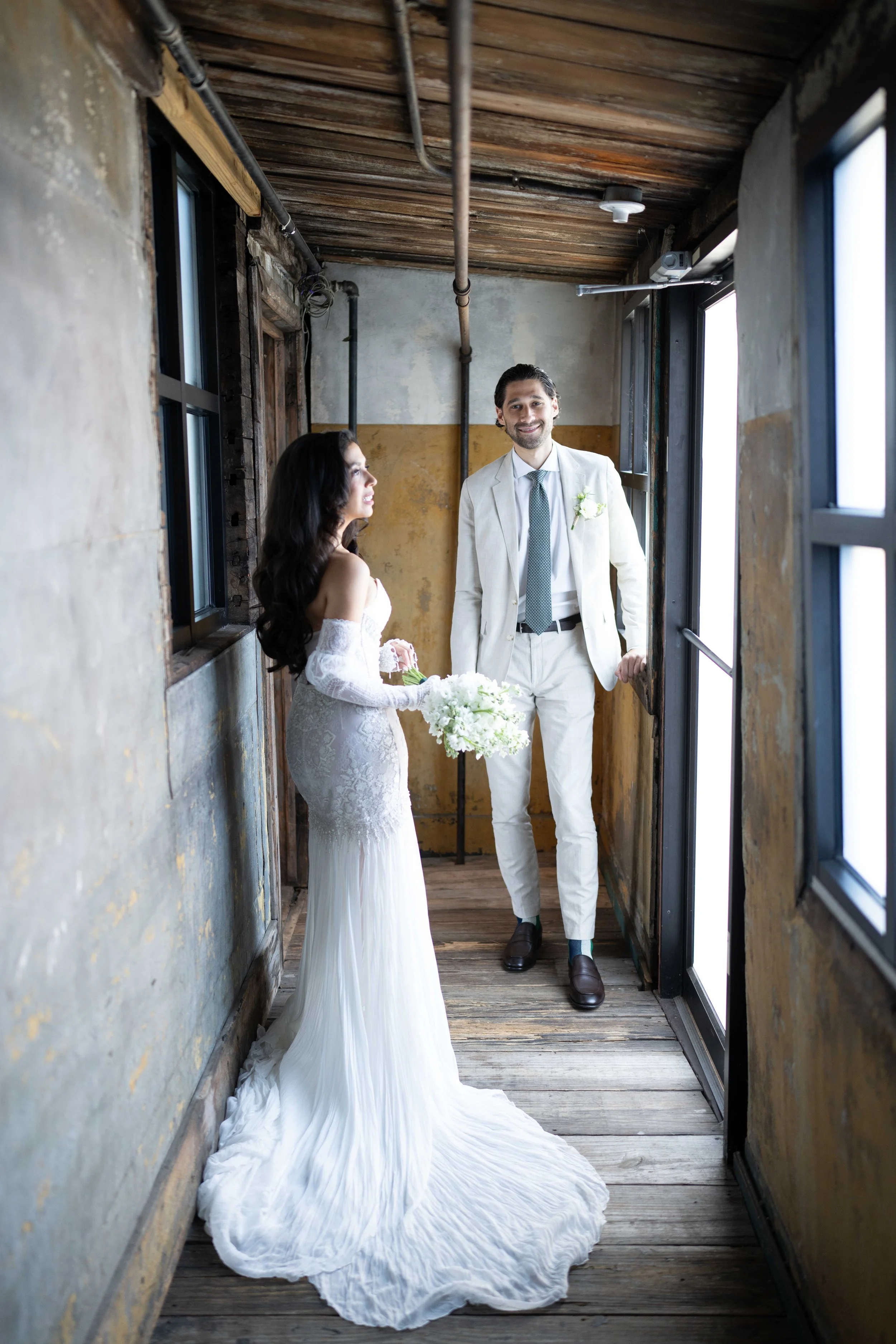A bride and groom standing in a rustic, industrial-style hallway with wooden floors and exposed pipes, smiling at each other.