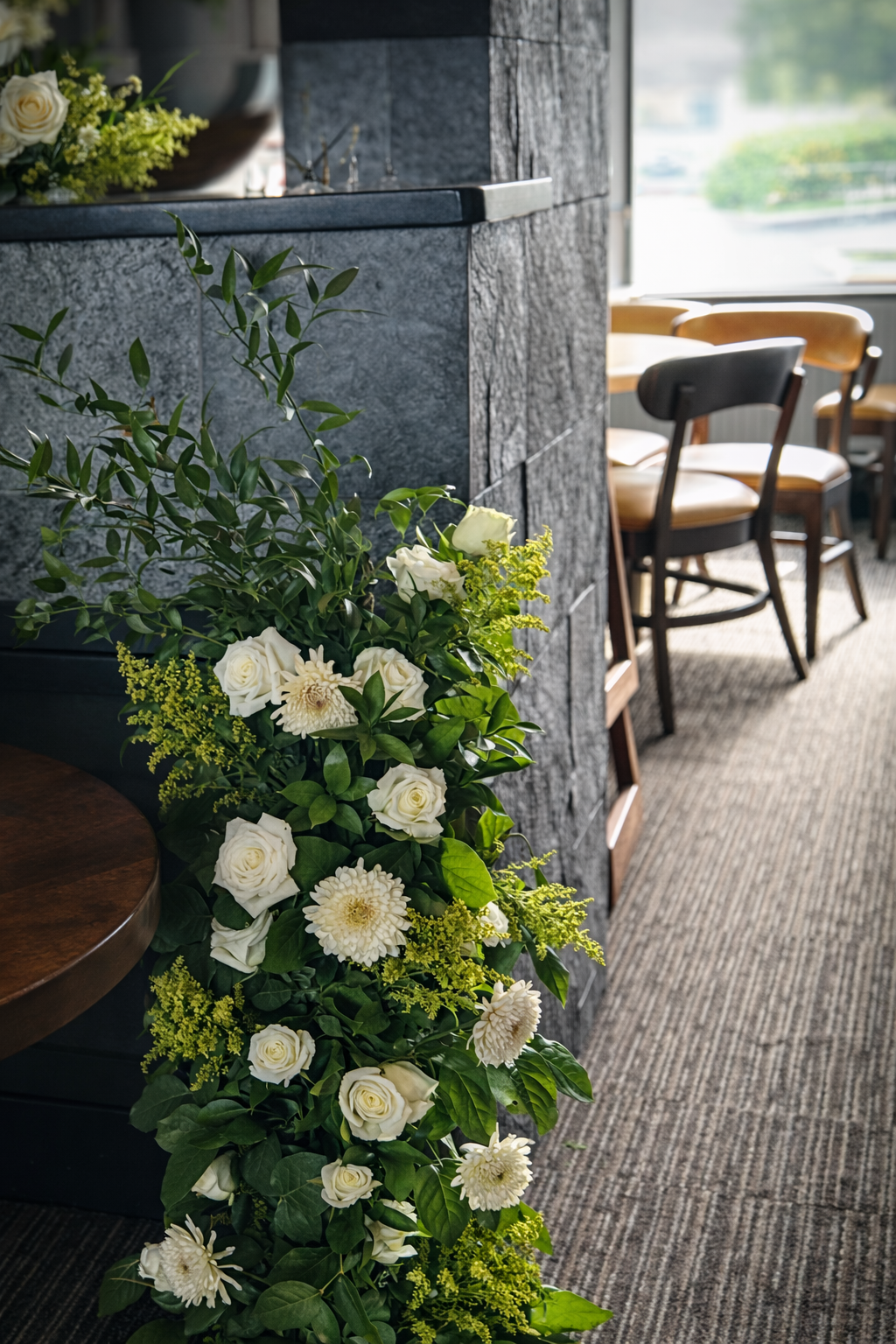 Indoor restaurant or cafe with a floral arrangement of white roses and greenery near a dark gray stone wall, with a wooden table and chairs in the background near a large window with a view of greenery outside.