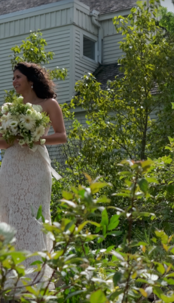 A woman in a white lace dress holding a bouquet of white flowers standing outdoors near a house and green trees.