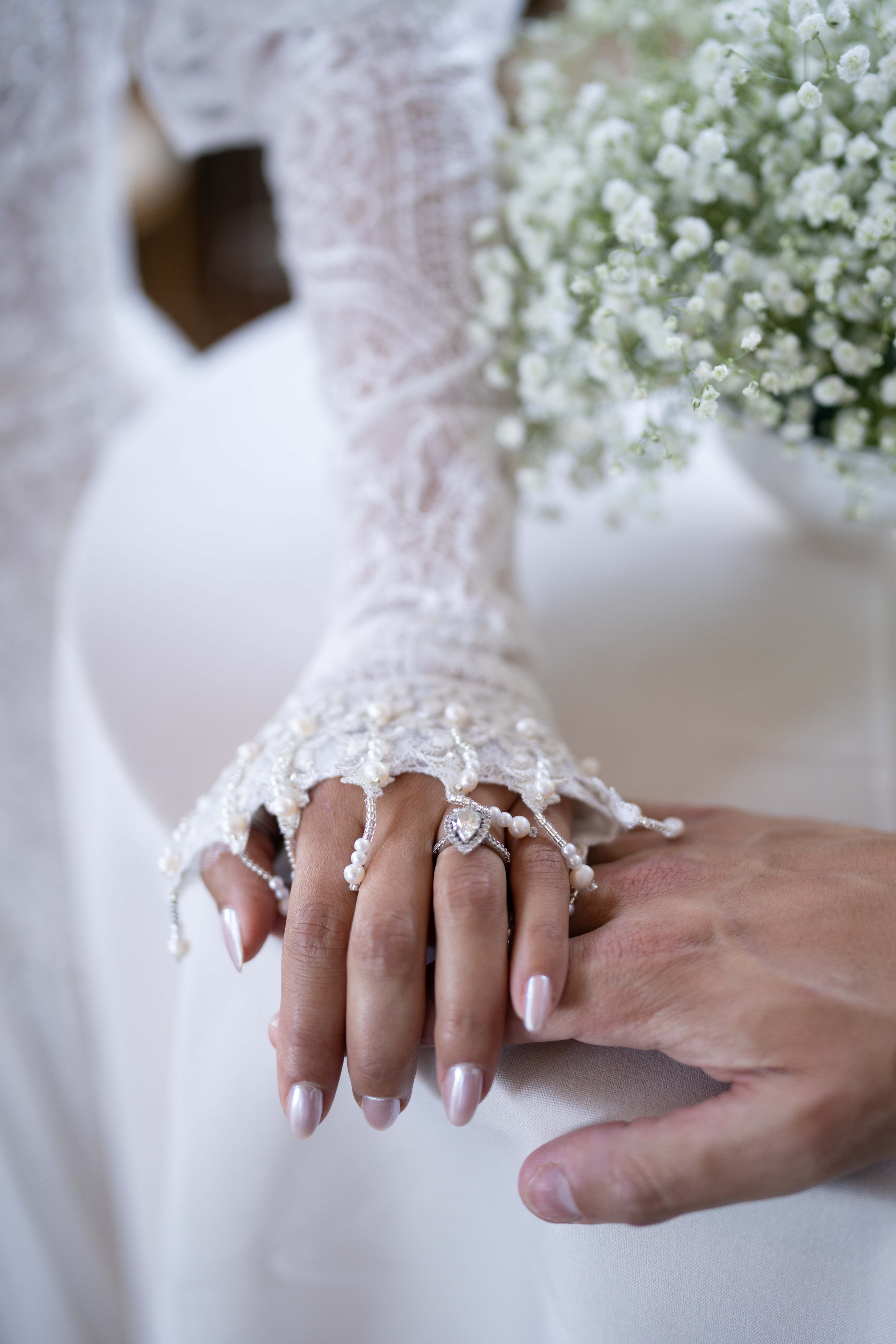 Close-up of a bride and groom holding hands, focusing on a pearl and diamond ring on the bride's finger. The bride's lace sleeve is decorated with pearls, and a bouquet of white baby's breath flowers is in the background.