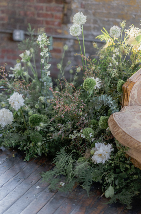 Indoor garden with various white and green flowering plants next to an ornate armchair on a wooden floor and brick wall background.