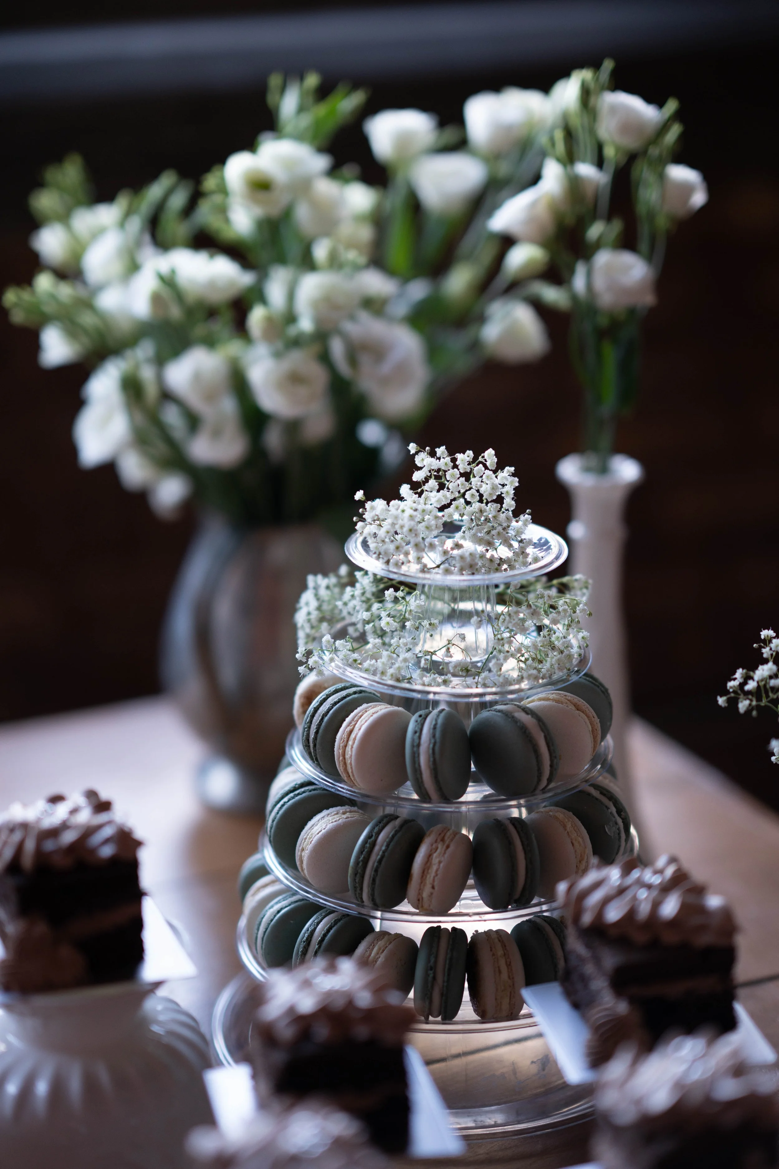 Elegant dessert display with a tiered stand of macarons and small floral arrangements, set against a background of white flowers in vases.