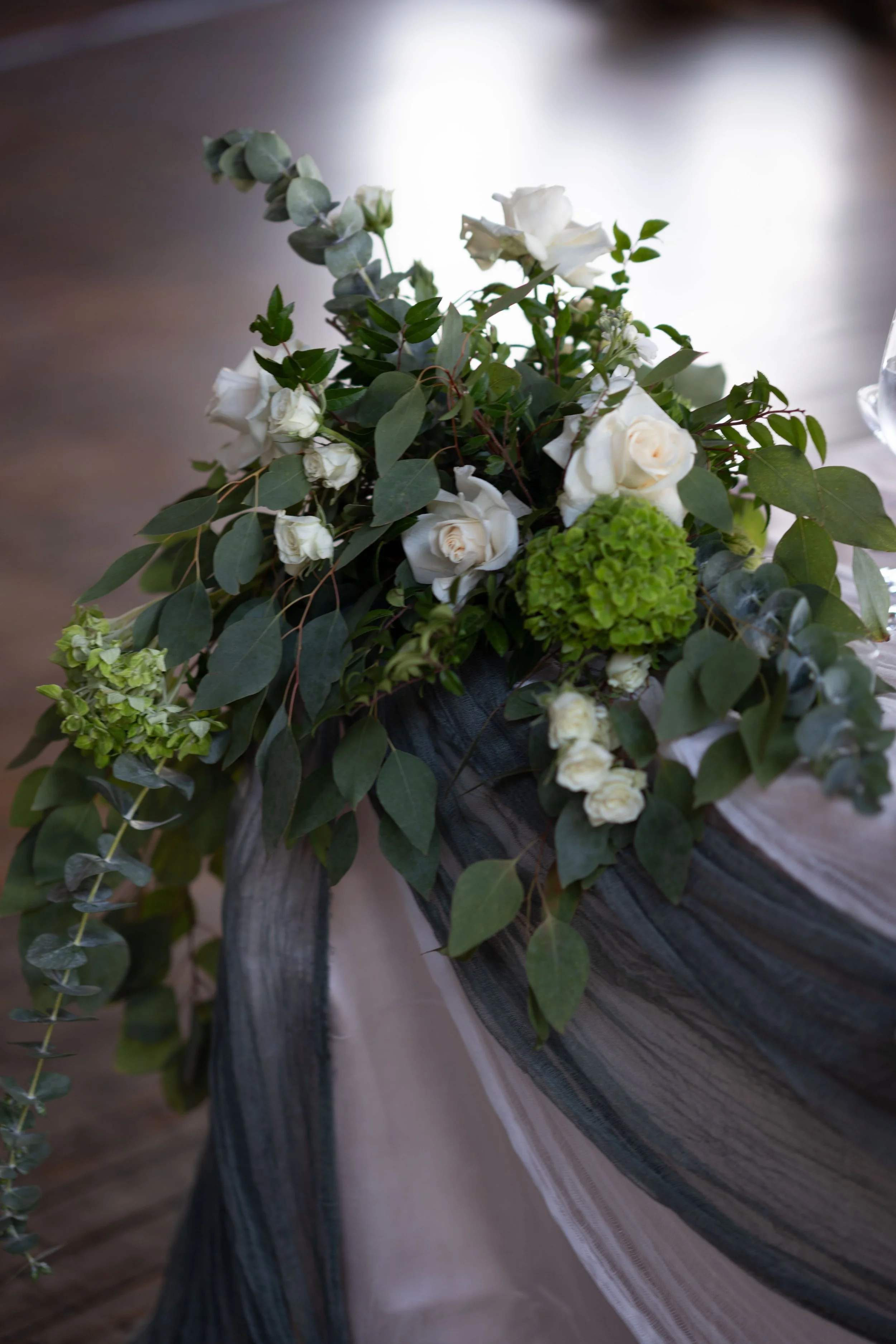 A floral arrangement with white roses, green hydrangea, eucalyptus leaves, and other greenery, placed on a table with a black and white tablecloth.