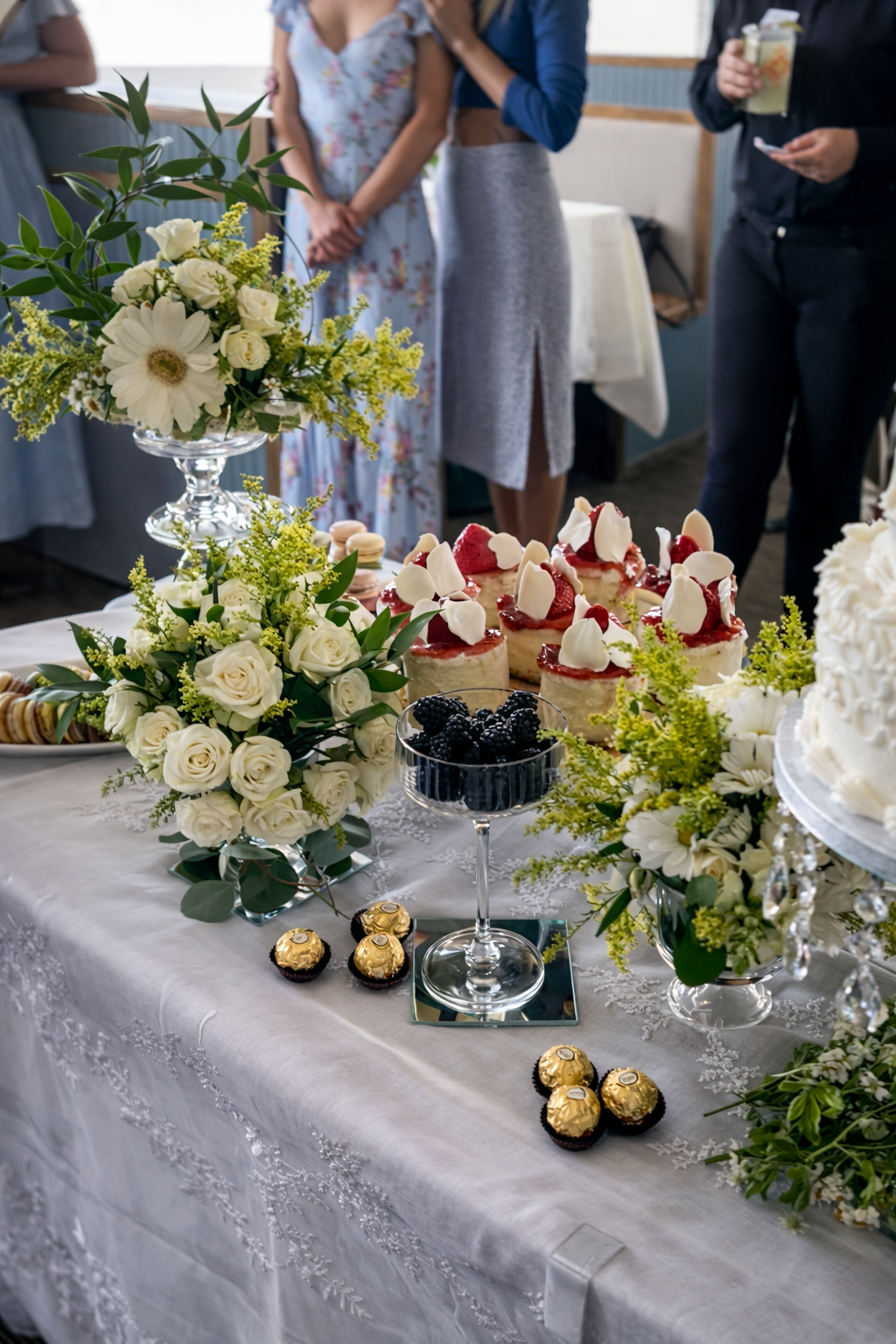 A table at a celebration, decorated with white floral arrangements and an assortment of desserts including strawberry shortcakes, a bowl of blackberries, and chocolates wrapped in gold foil. People are standing in the background, some holding drinks.