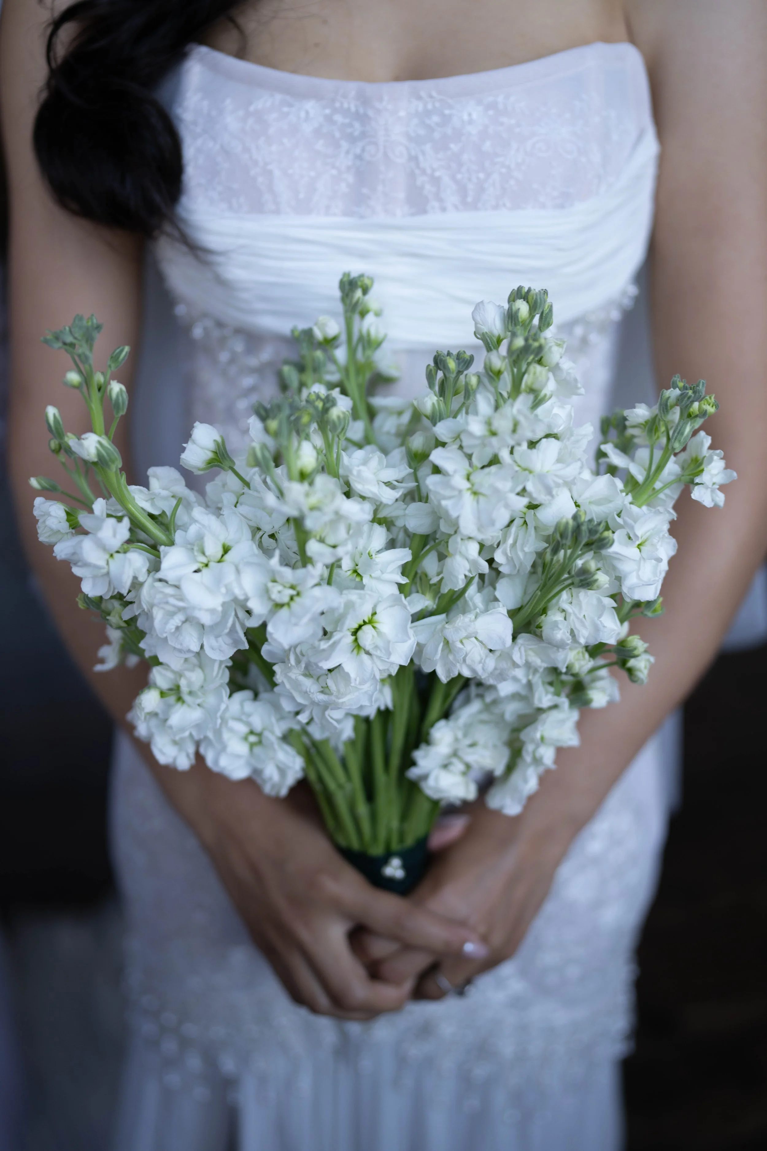 A bride holding a bouquet of white flowers, wearing a white dress with lace and sheer fabric details.