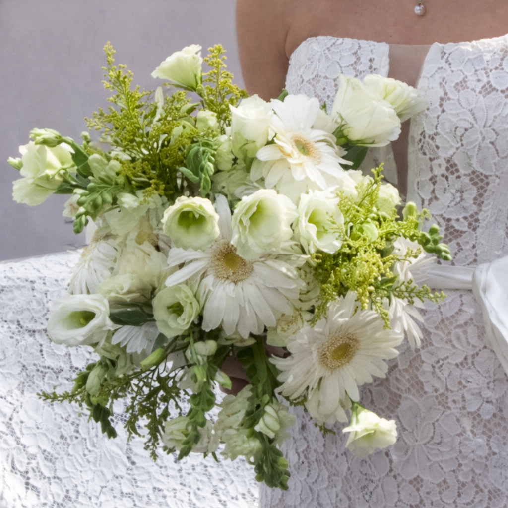 A bride holding a bouquet of white and cream flowers including daisies, roses, and greenery, against a neutral background and wearing a white lace dress.