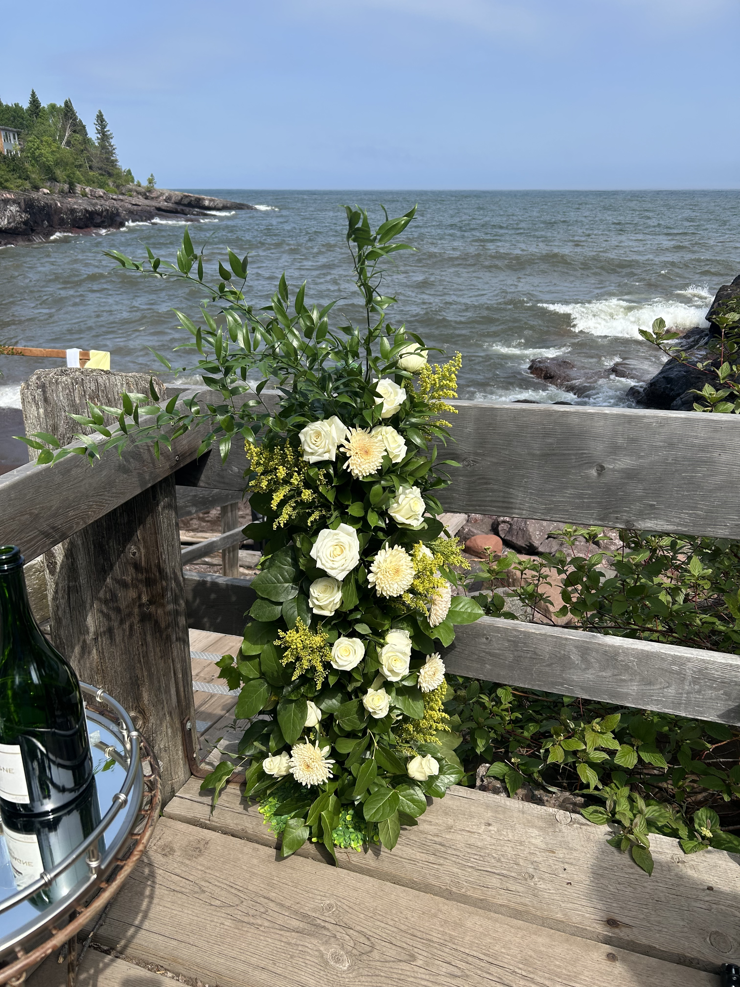 A floral arrangement with white roses, cream-colored chrysanthemums, and greenery placed against a wooden railing on a deck overlooking a lake with rocky shoreline and waves.