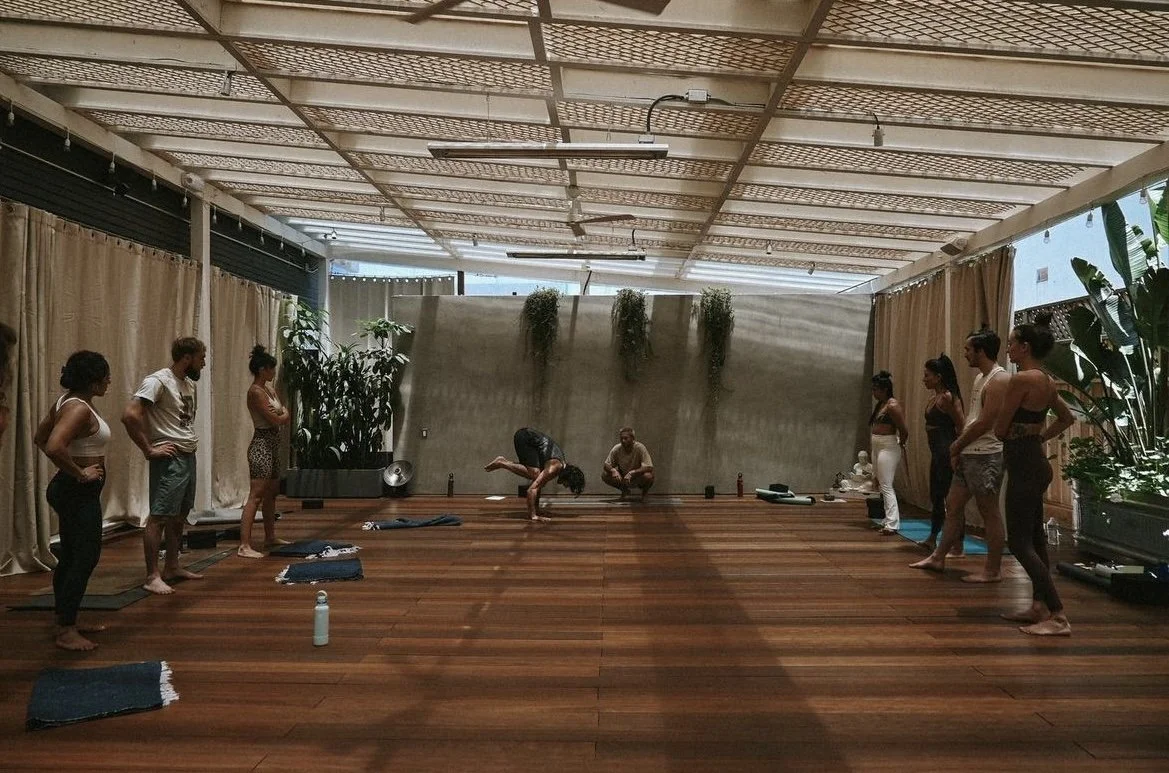 People participating in a yoga class indoors, with mats on the wooden floor, some sitting and some standing, in a spacious room with plants and curtains.