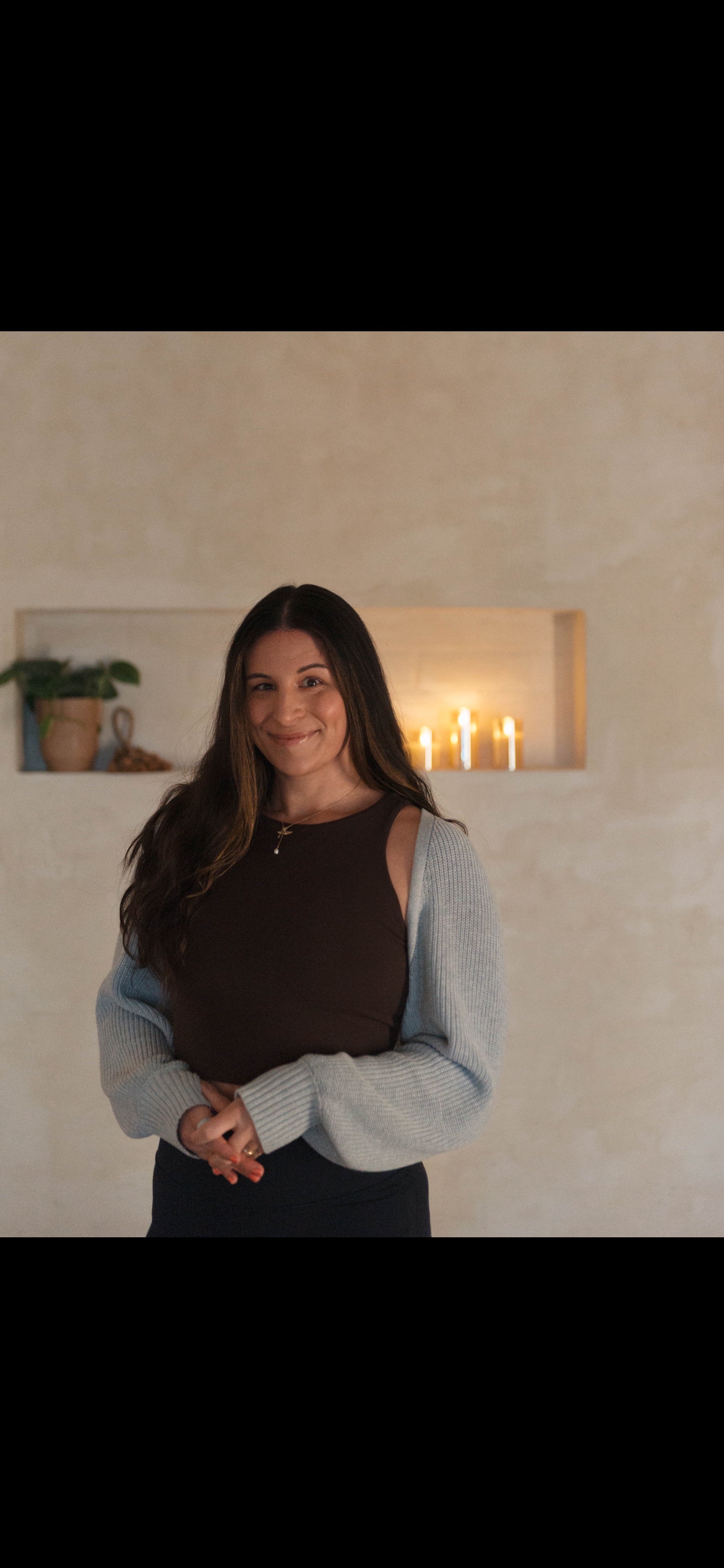 A woman with long dark hair smiling and standing in a softly lit room with candles and a small shelf with potted plants in the background.