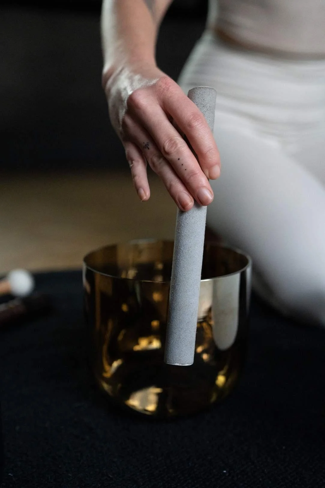 Close-up of a hand with small tattoos holding a gray stick over a golden reflective bowl on a black surface, with a person's knee and white pants in the background.