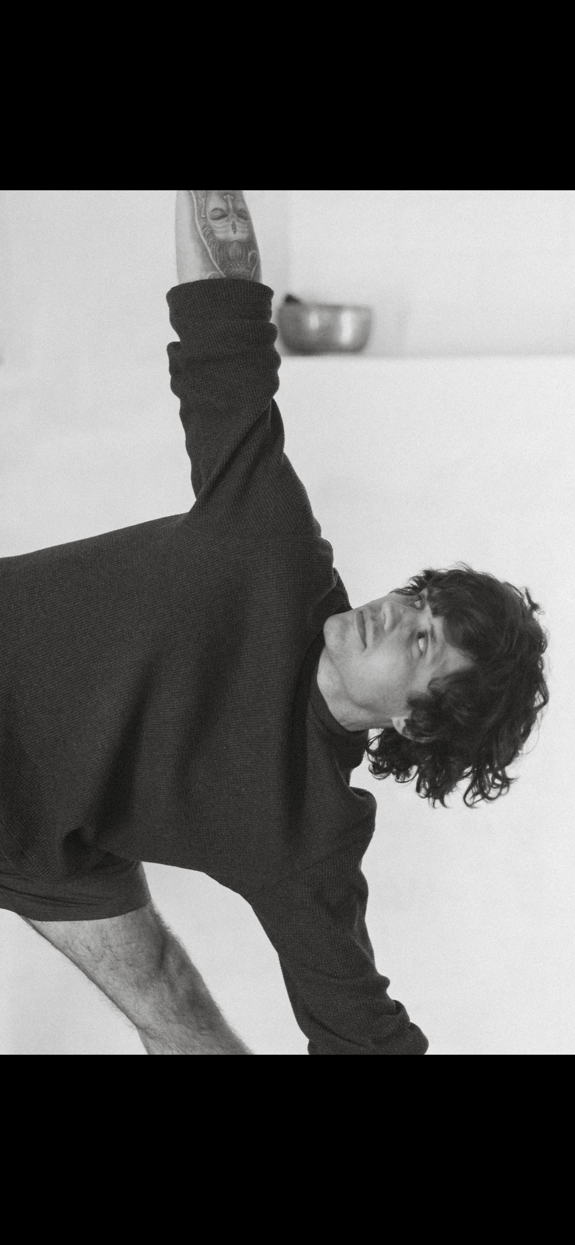 Black and white photo of a young man with curly hair, lying down with one arm raised and bent at the elbow, wearing a dark sweater, with a tattoo on his arm, looking upward in a minimalist room with a white wall and a metal bowl in the background.