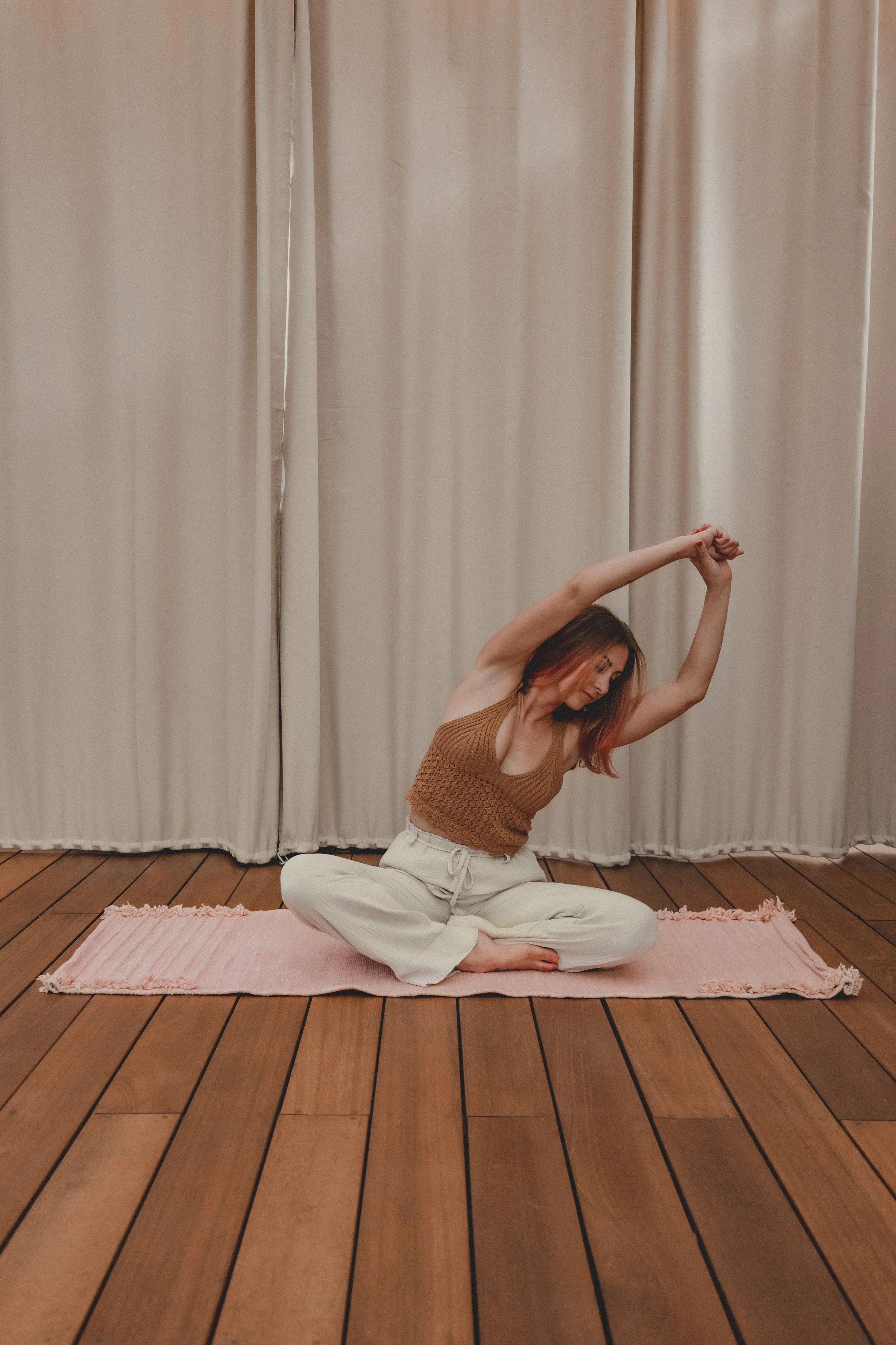 Woman doing yoga stretch in a room with wooden floor and beige curtains, sitting cross-legged on a pink mat.