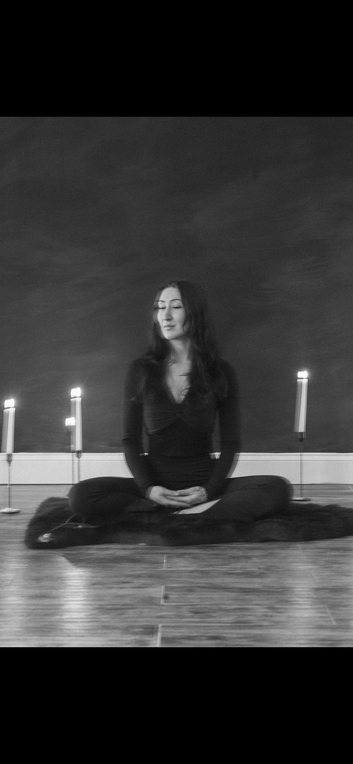 A woman sitting cross-legged on a rug, with closed eyes, surrounded by four lit candles in a dark room.
