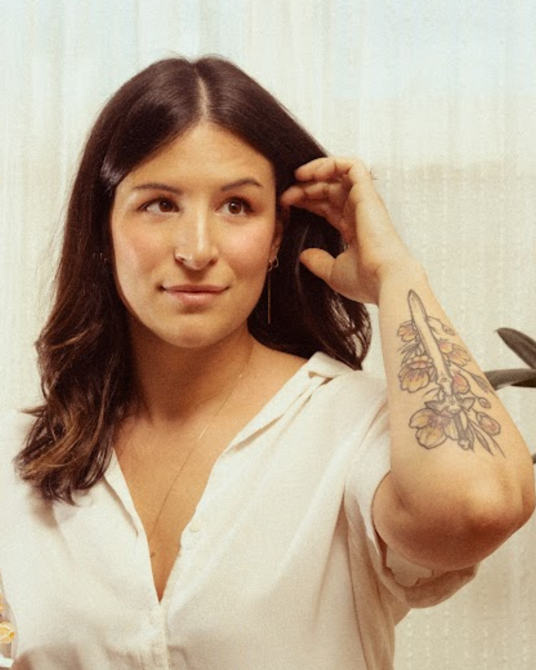 A woman with brown hair and a tattoo on her forearm, touching her hair and looking slightly to her right, wearing a cream-colored blouse and jewelry, against a plain light-colored background.
