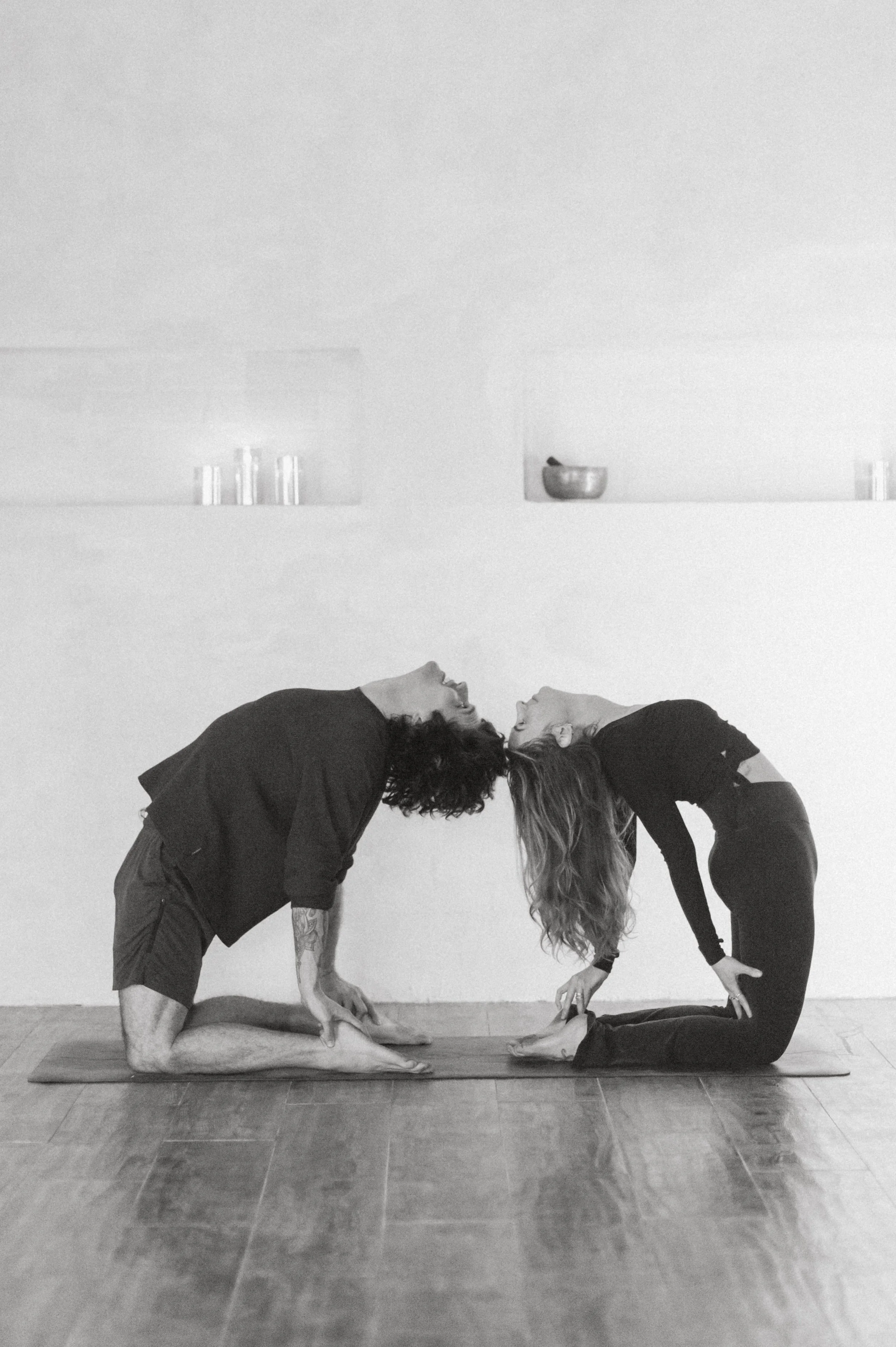 Two women and a man practicing yoga together in a studio. They are performing a pose on a yoga mat, leaning forward with their heads touching and facing each other, in a mirrored pose. The background shows a minimalistic wall with a few shelves holding glasses, a bowl, and cans.