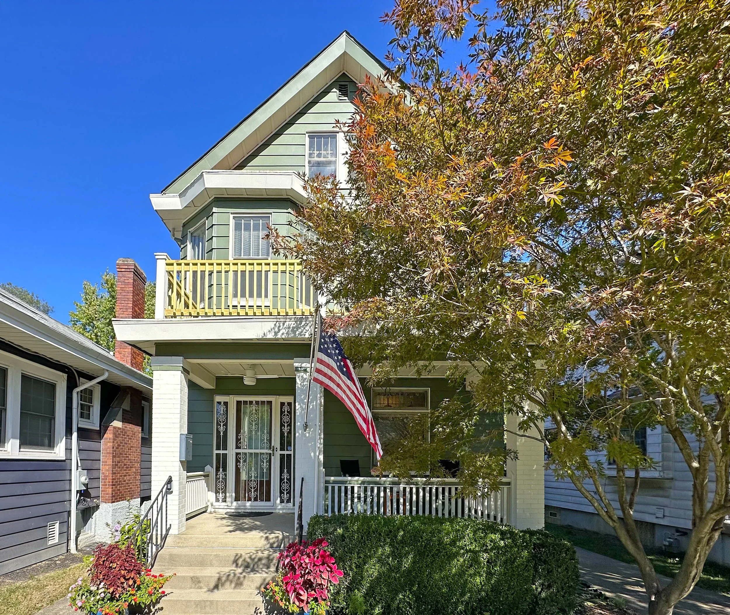 Front view of a three-story house with a green exterior, white trim, and a second-floor balcony with a yellow railing. An American flag hangs near the front door, which is accessed by stairs. The yard has a manicured bush and pink flowers, with a mature tree on the right side and neighboring houses visible.