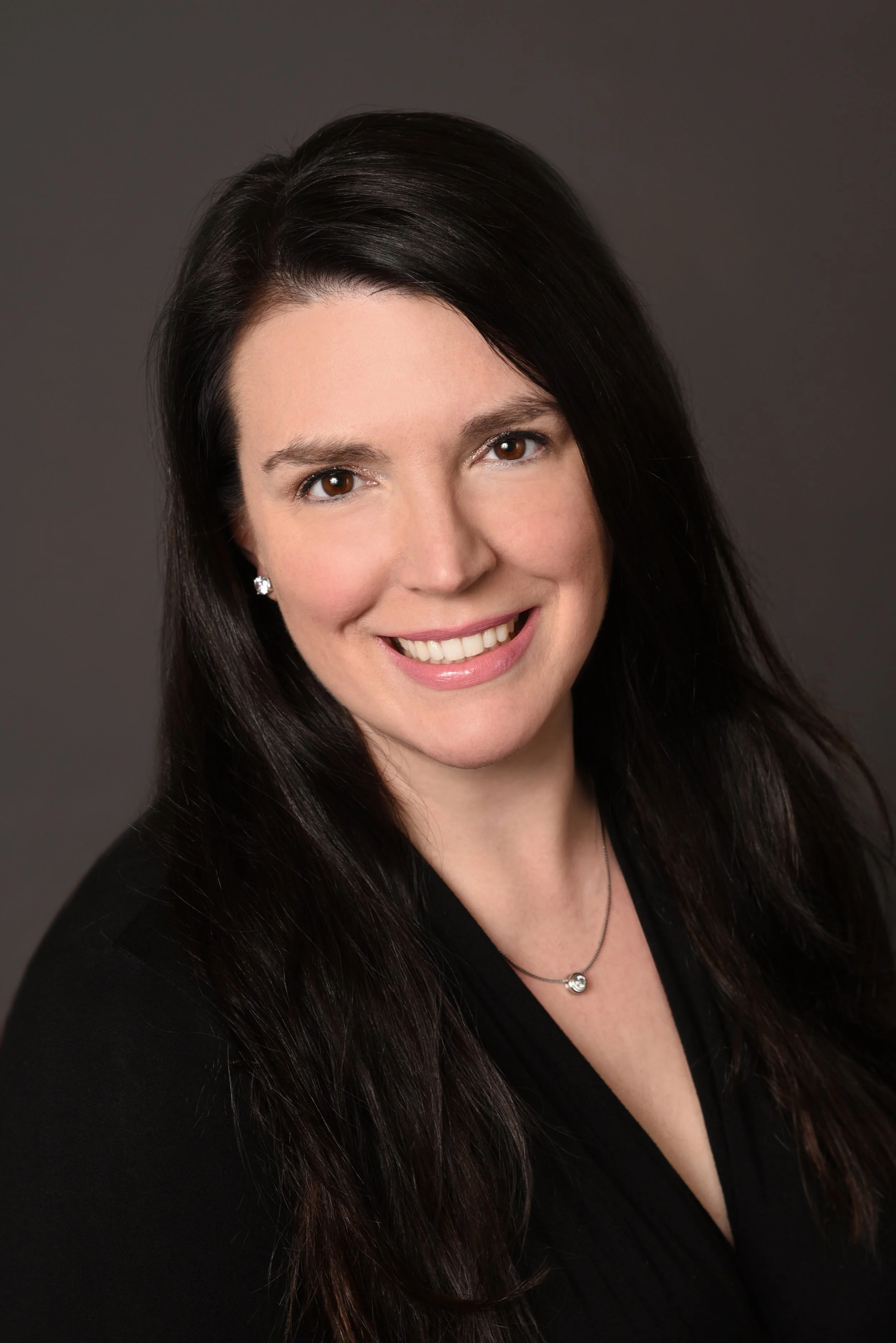 Professional headshot of a woman with long dark hair, smiling, wearing a black top, earrings, and a necklace, against a dark background.