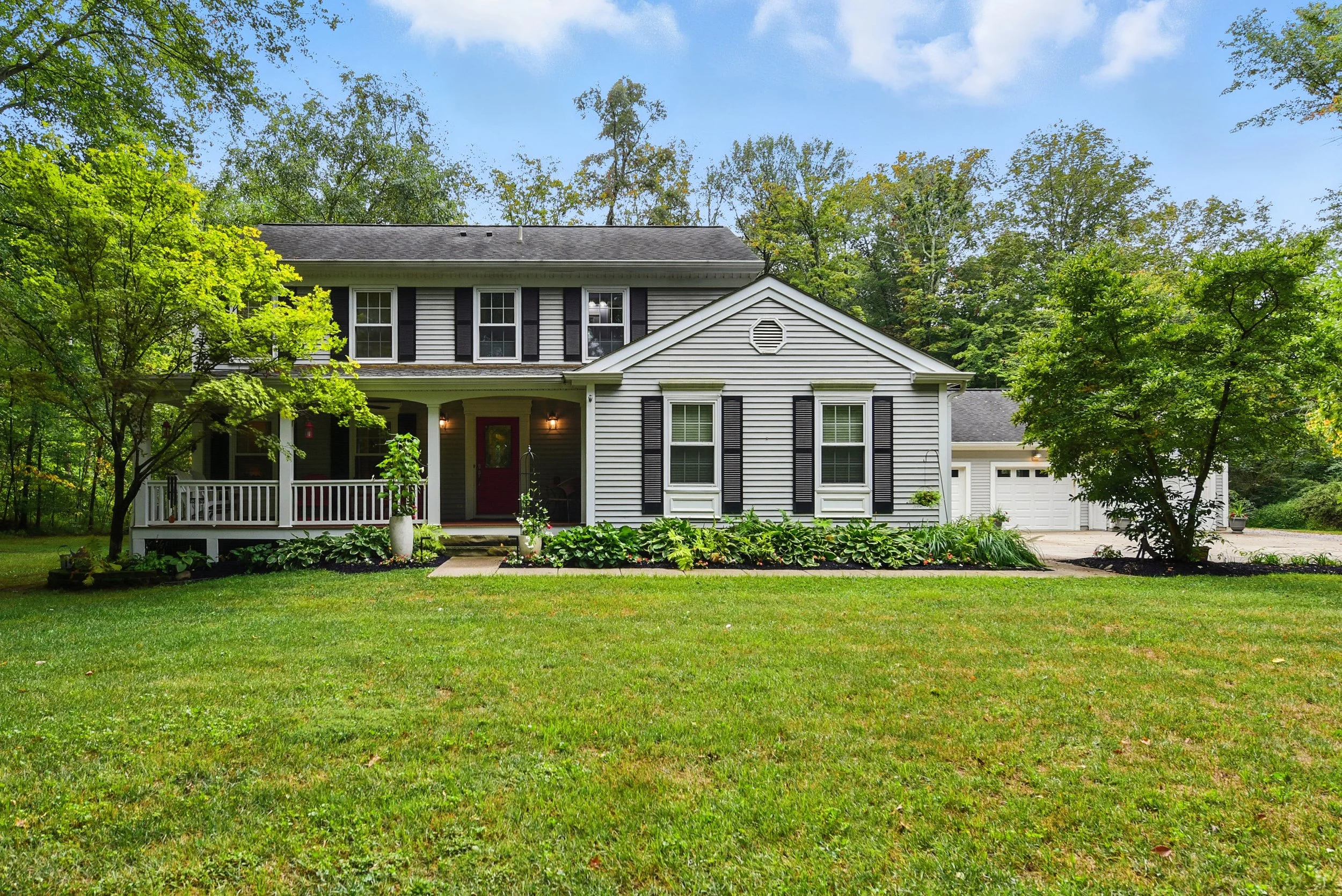 Front view of a two-story house with white siding, black shutters, and a red front door, surrounded by green trees and a well-maintained lawn.