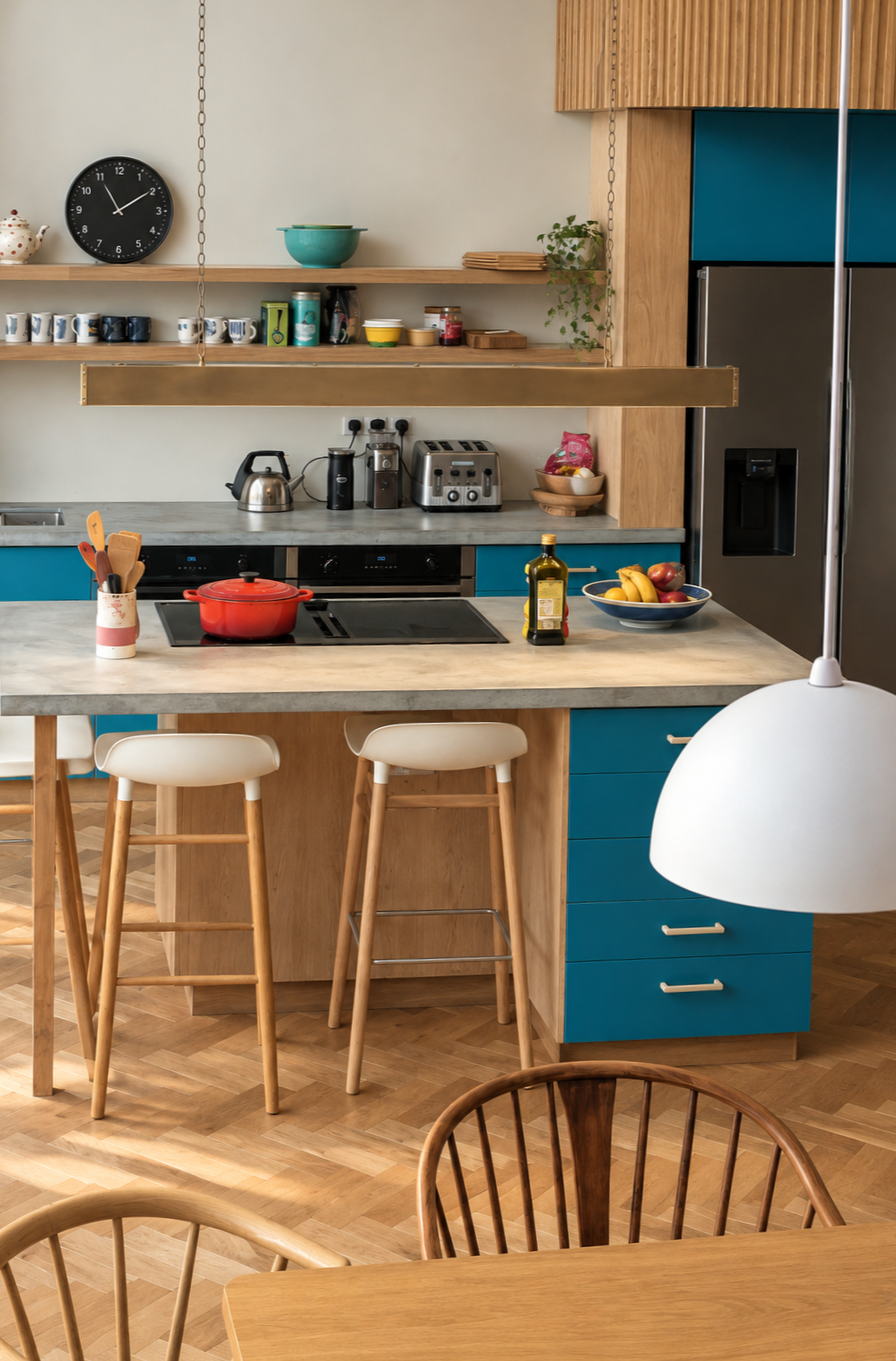 Modern kitchen with wooden cabinetry, blue drawers, a marble countertop, and kitchen appliances including a toaster, kettle, and refrigerator. A bowl of bananas and apples, a bottle of olive oil, and a red pot on the stove are visible. There are two barstools and dining chairs in the foreground.