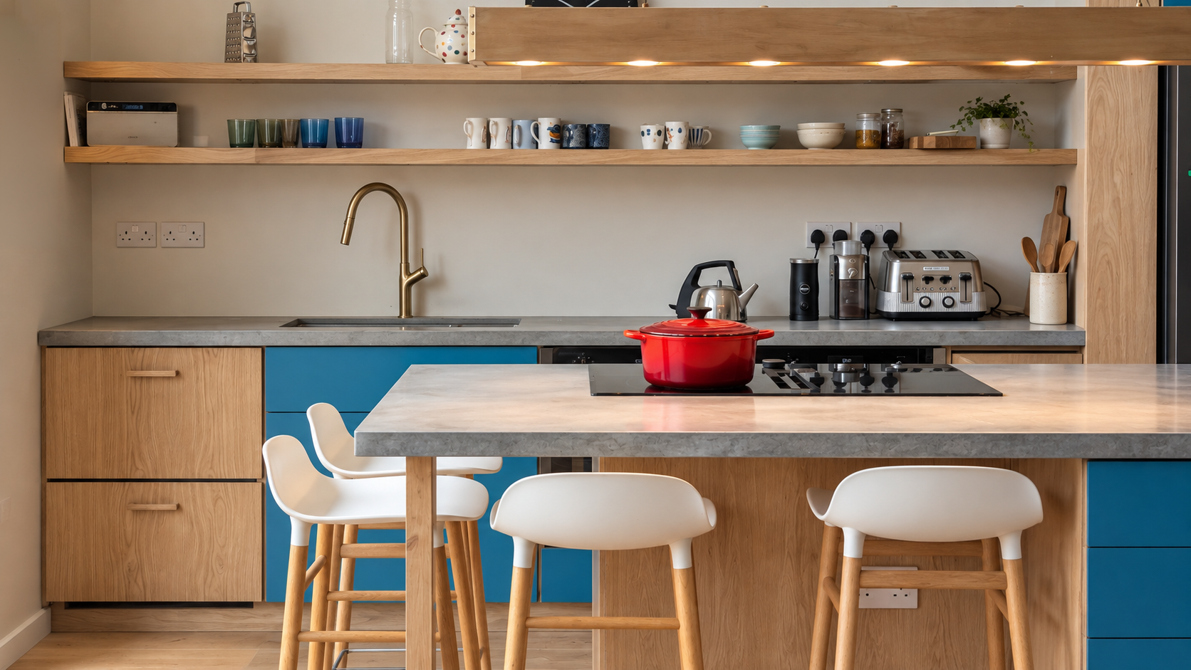 Modern kitchen with open wooden shelves, a beige wall, a marble countertop, and a blue and wood-colored kitchen island. There are white and blue stools at the island, a red pot on the stove, and various kitchen appliances and mugs on the counter and shelves.
