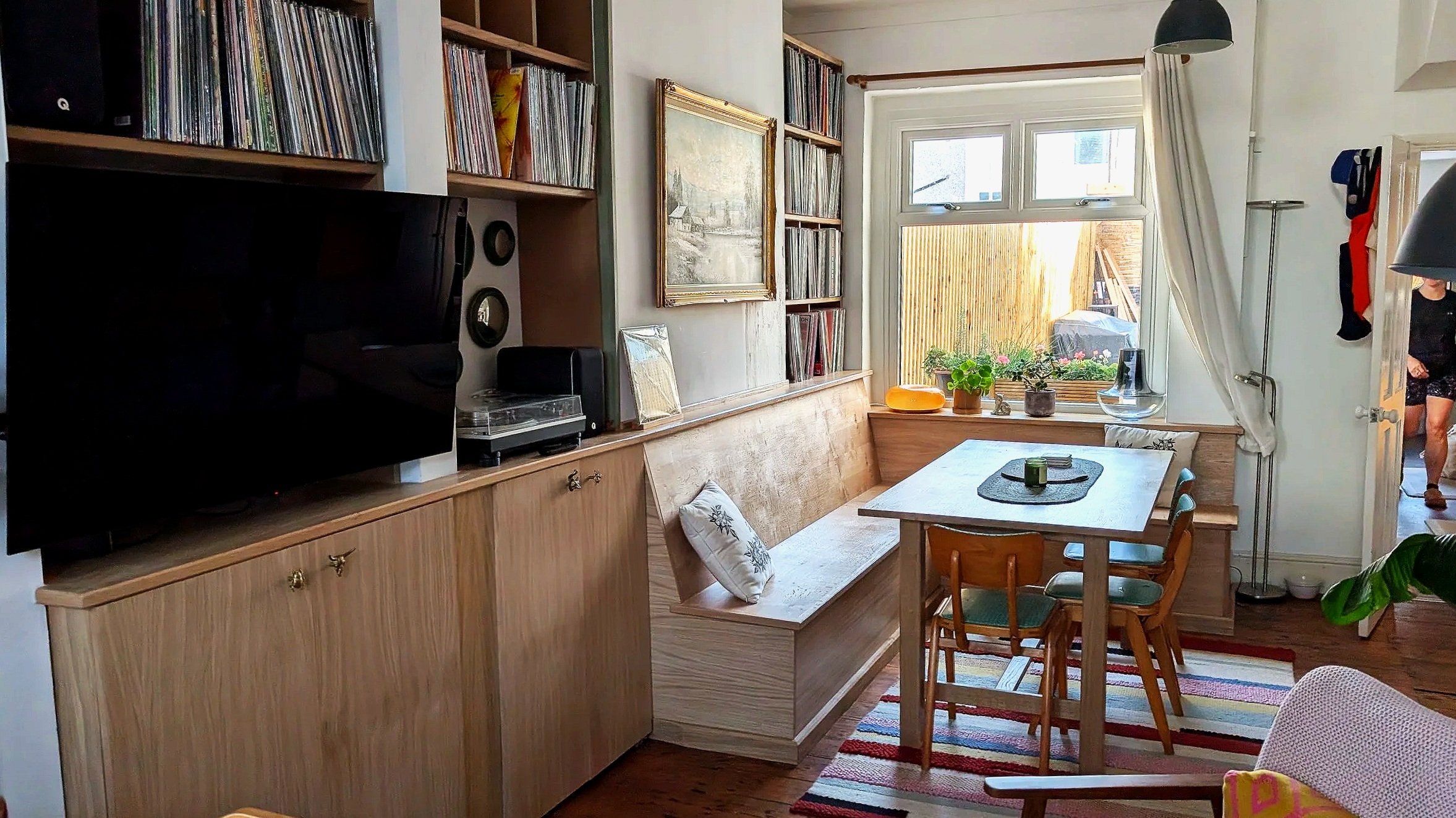 A cozy dining area with a wooden table and chairs near a large window, decorated with potted plants, and surrounded by bookshelves filled with books and vinyl records.
