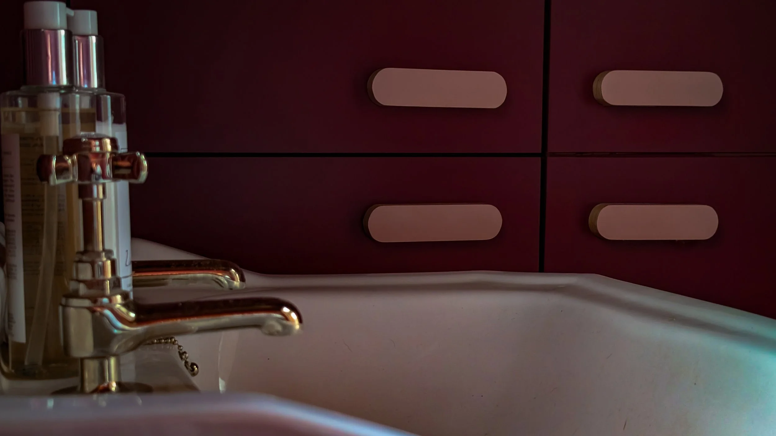 Close-up of a bathroom sink with a chrome faucet and two clear soap dispensers on the side, set against a dark red cabinet with four doors with intricately designed and handcrafted bespoke handles by Scribe Design Studio.