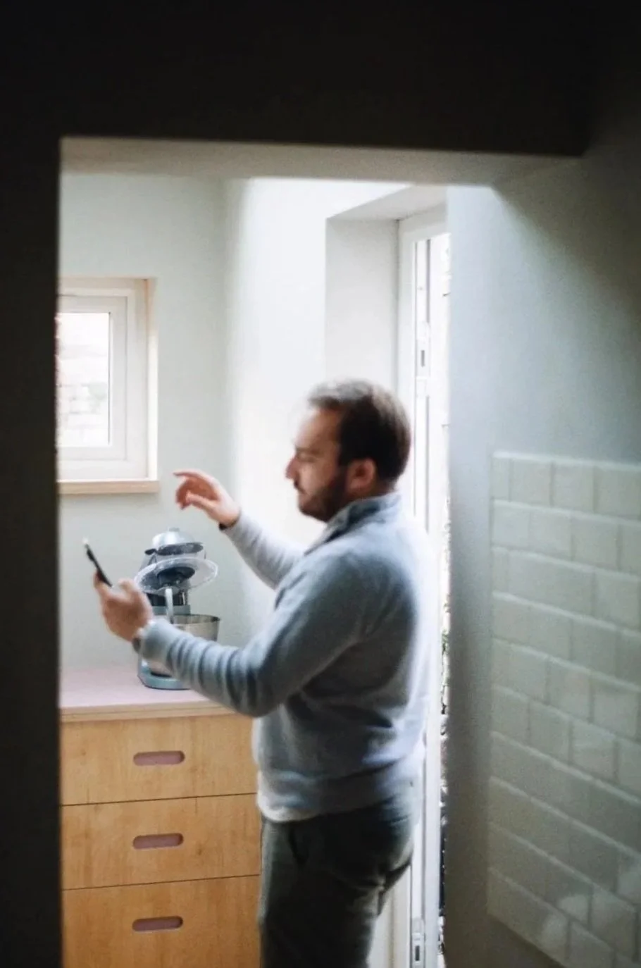 Reuben Dutch Interior–Spatial Designer project managing a project - standing in a kitchen, looking at his phone, with a birch plywood counter top and a coffee maker visible behind him.