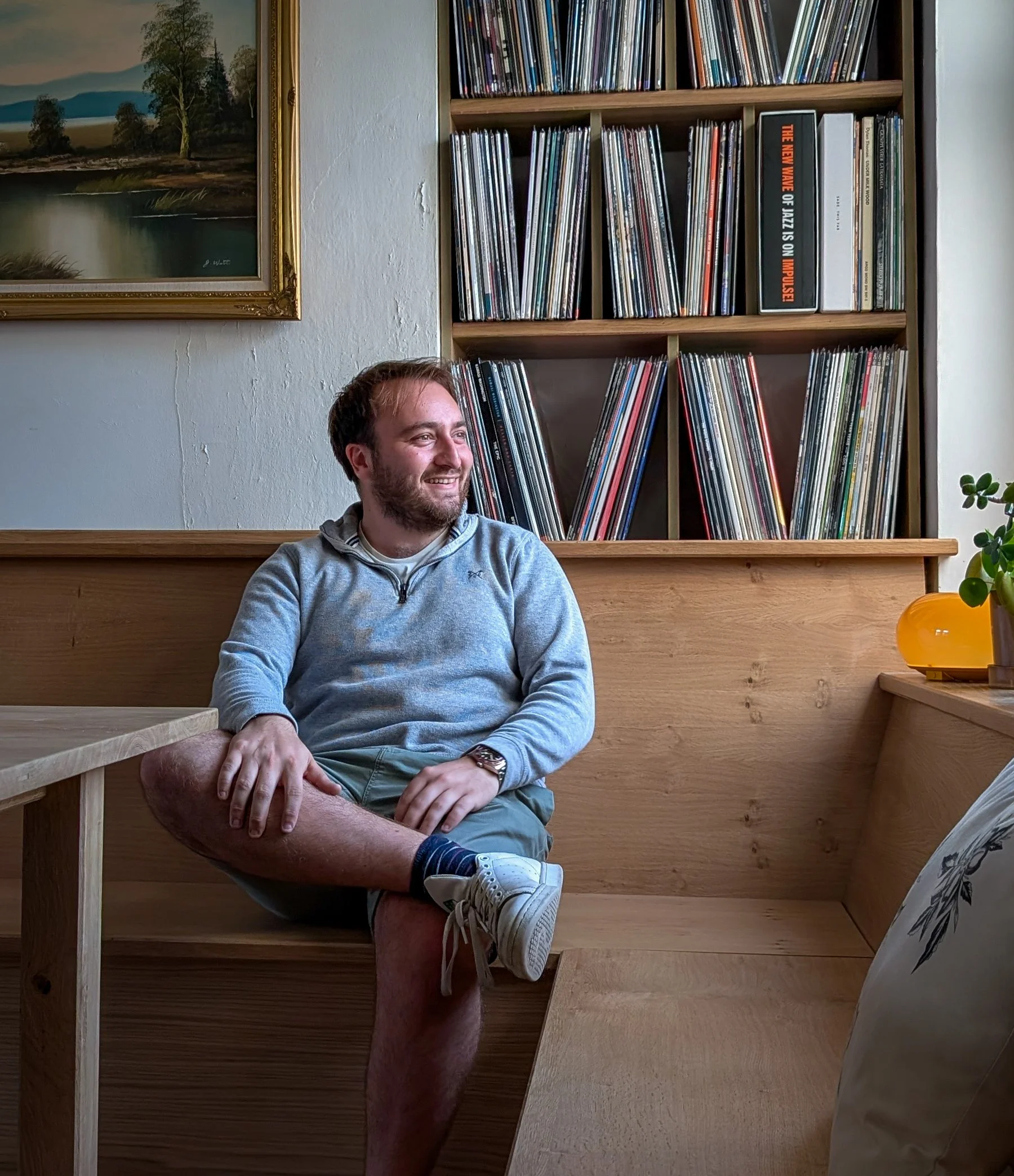 Reuben Dutch Interior–Spatial Designer seated at a wooden table, smiling and looking to the left. Behind him is a bespoke record shelf filled with vinyl records and a landscape painting on the wall.