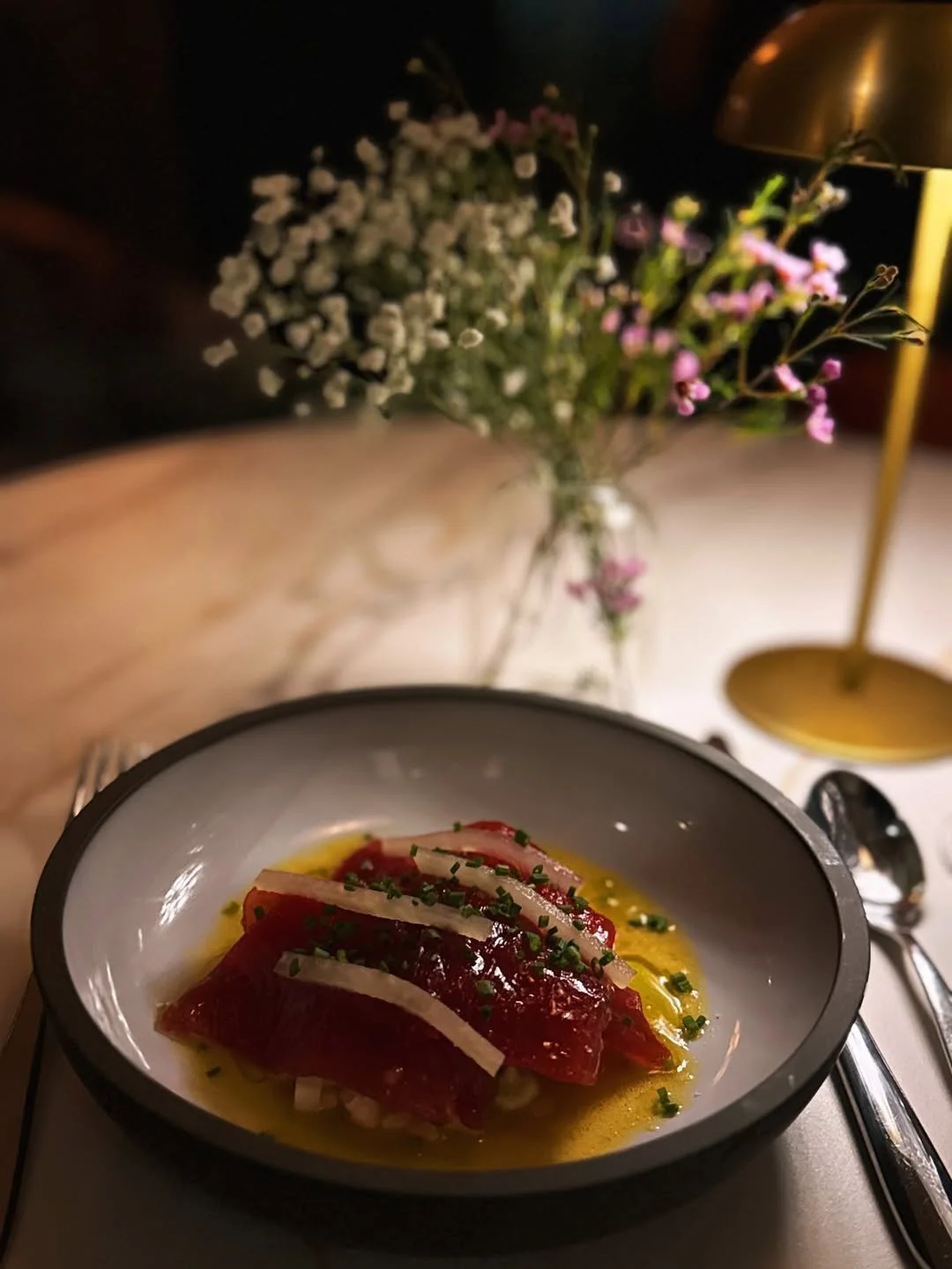 A close-up, dimly lit shot of a thinly sliced red crudo or carpaccio dish garnished with chives and white radish slivers, served in a shallow grey bowl with a vibrant yellow citrus oil base. In the background, a small vase of delicate white and pink 