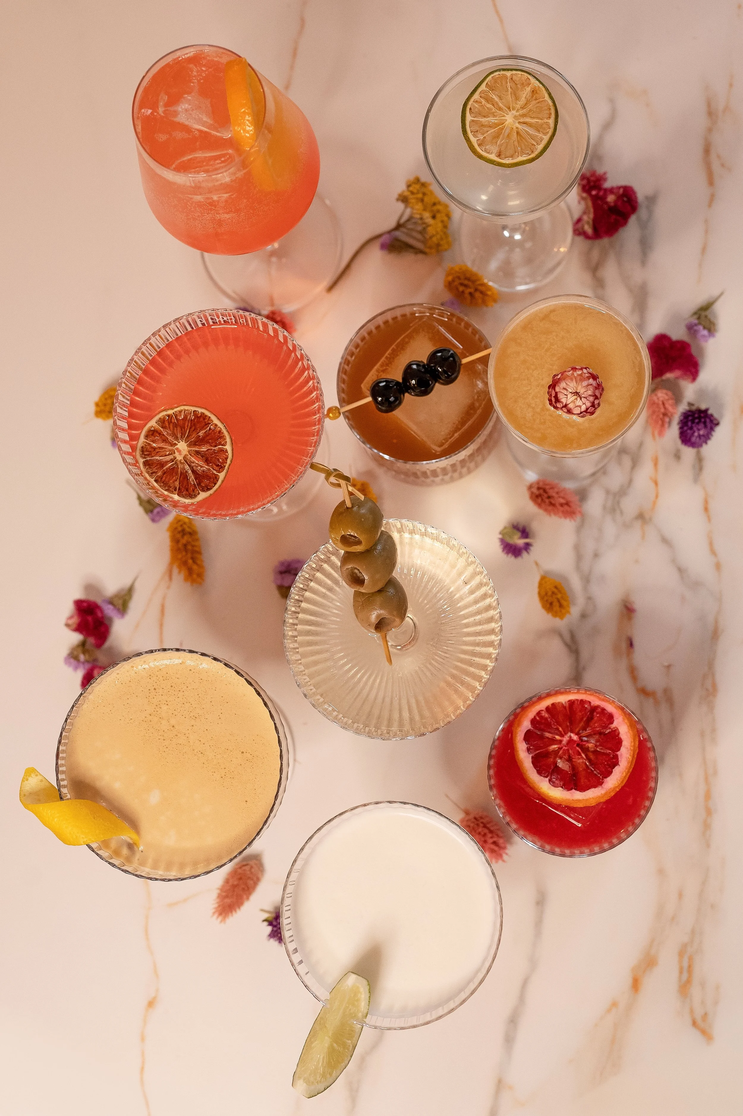 Flatlay view of various craft cocktails at Viv & Jules, showcasing colorful garnishes and premium glassware on a marble tabletop.