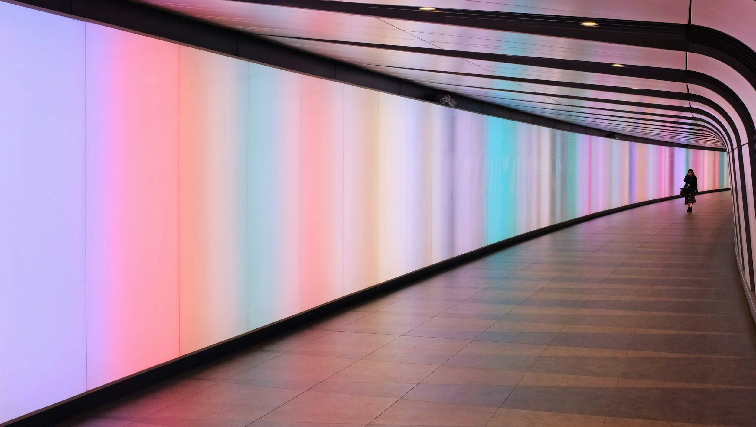 An illuminated multicolored LED wall in an urban tunnel with a lone person walking in the distance.
