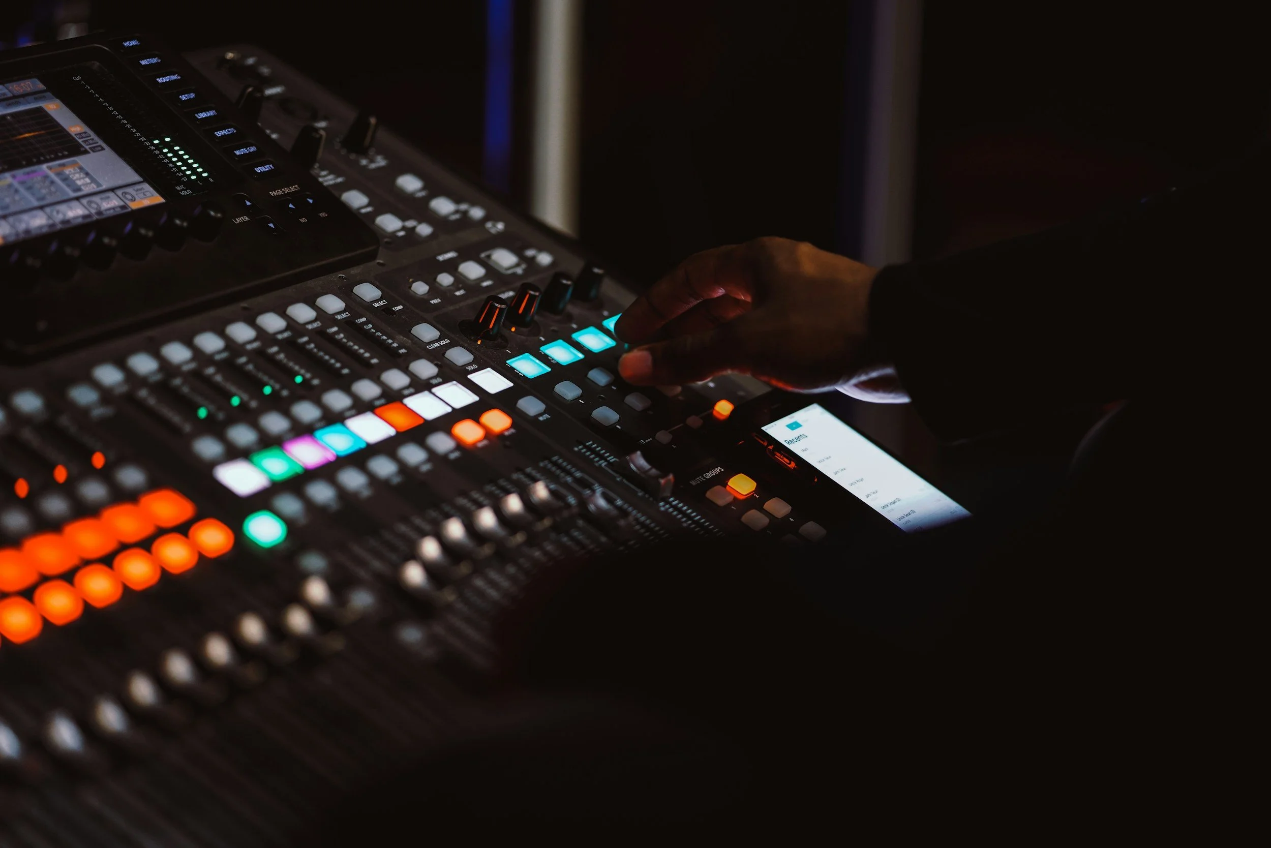 Close-up of a person's hand operating a professional digital audio mixing console with colorful illuminated buttons and sliders, in a dark environment.