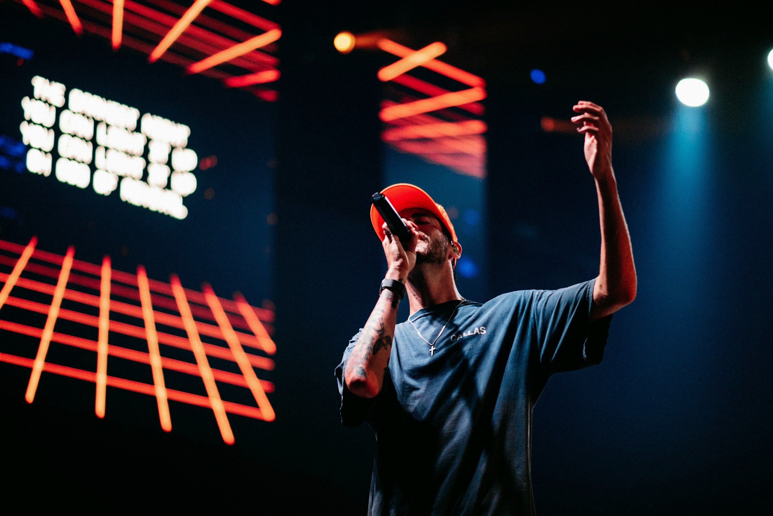 A man wearing a red baseball cap and a dark T-shirt with a cross necklace, holding a microphone, performing on stage with colorful lights and LED screens in the background.