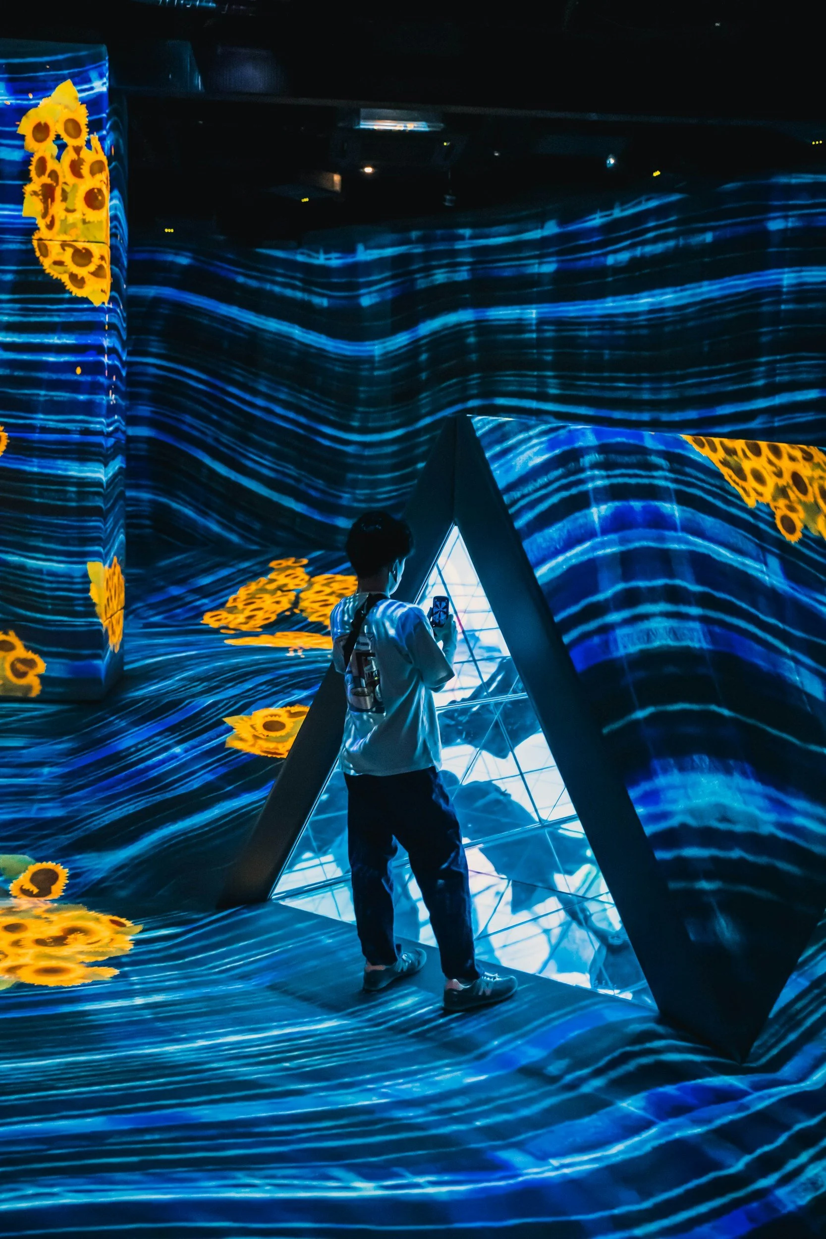 Person standing in an immersive digital art installation with blue wavy patterns and yellow sunflower-like designs projected onto the walls and floor, taking a photo with a smartphone.