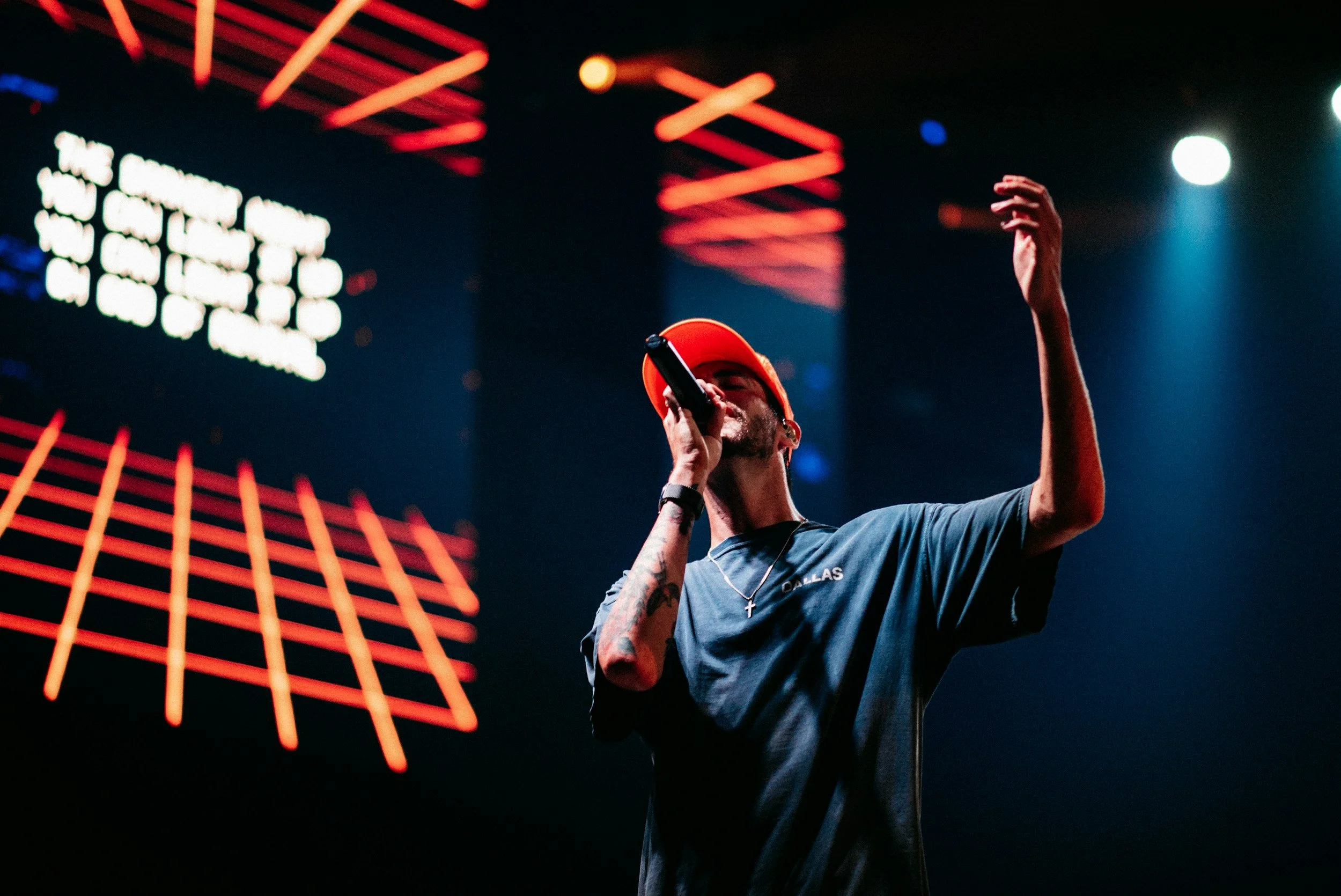 A man wearing an orange cap and a dark T-shirt with 'DALLAS' written on it is singing into a microphone on stage, with one arm raised. The background features stage lights and a digital display with bright lights.