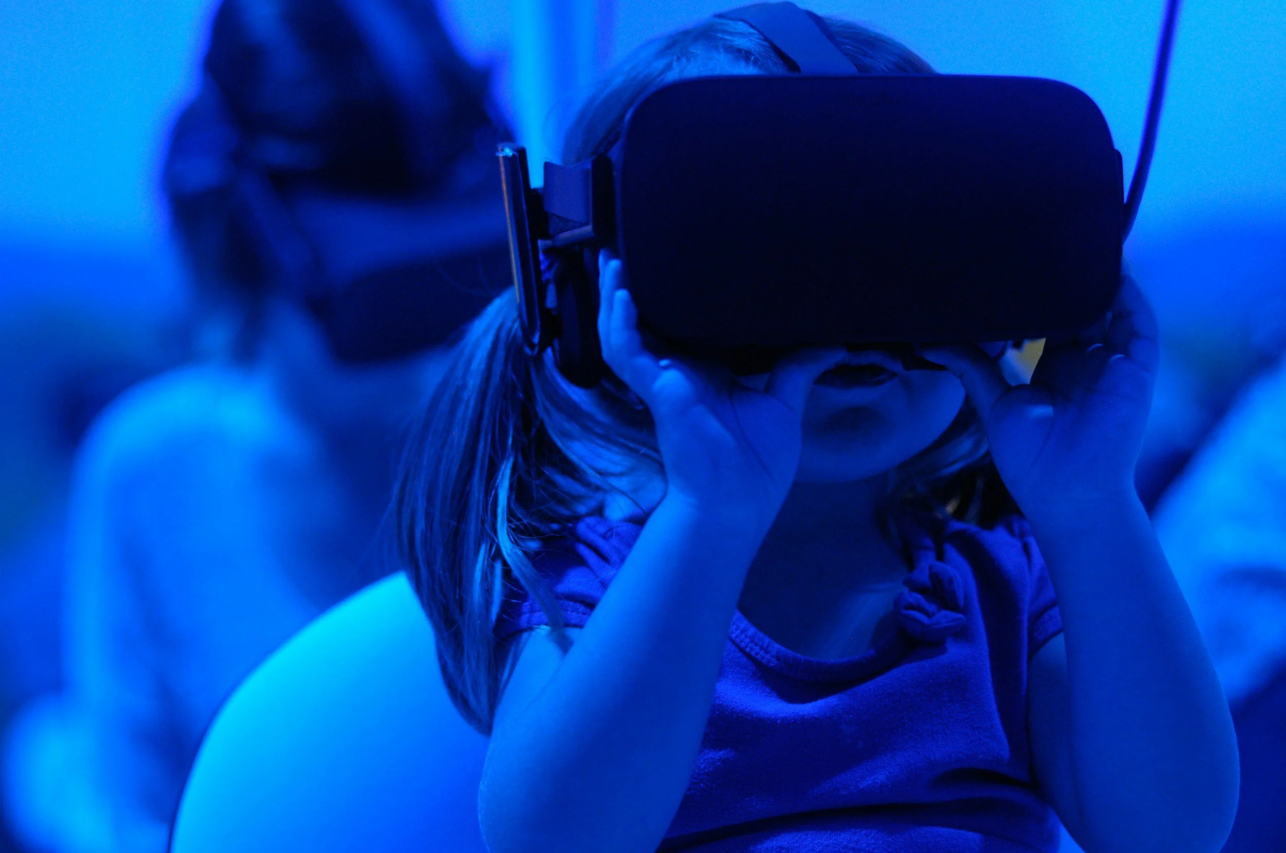 A young girl wearing virtual reality goggles, holding them with both hands, in a room illuminated with blue light.
