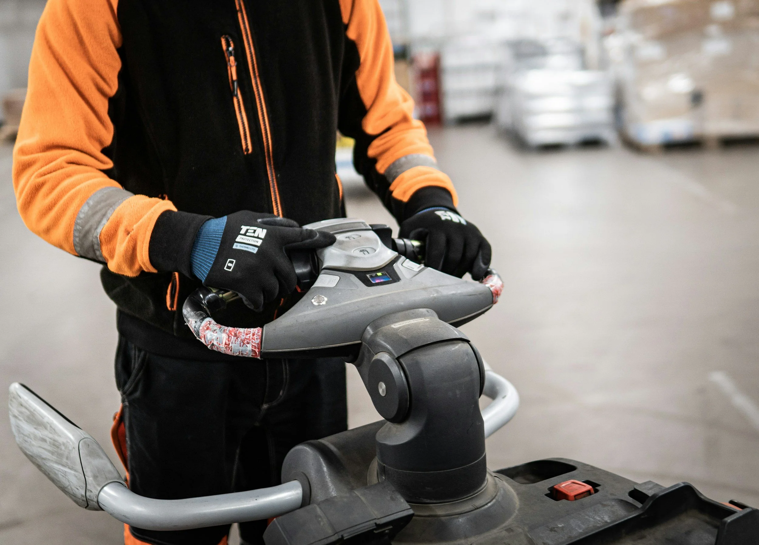 Person in black and orange jacket and black gloves operating a floor polishing or buffing machine in a warehouse.