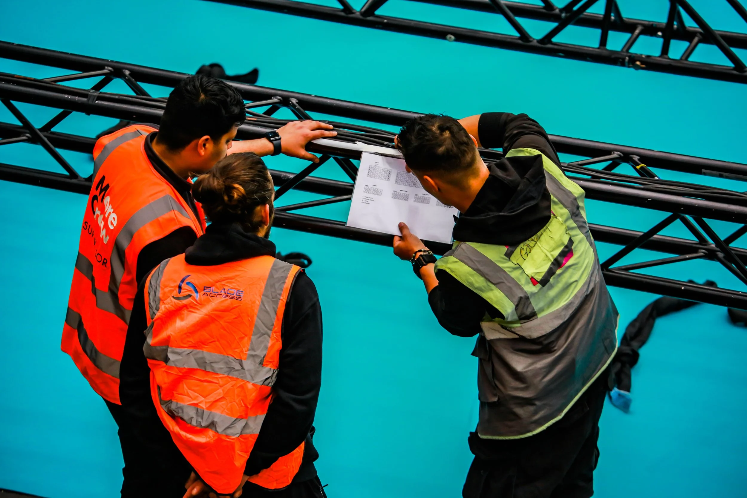 Three event staff members wearing orange safety vests working together on a black metal truss structure with blue flooring in the background.