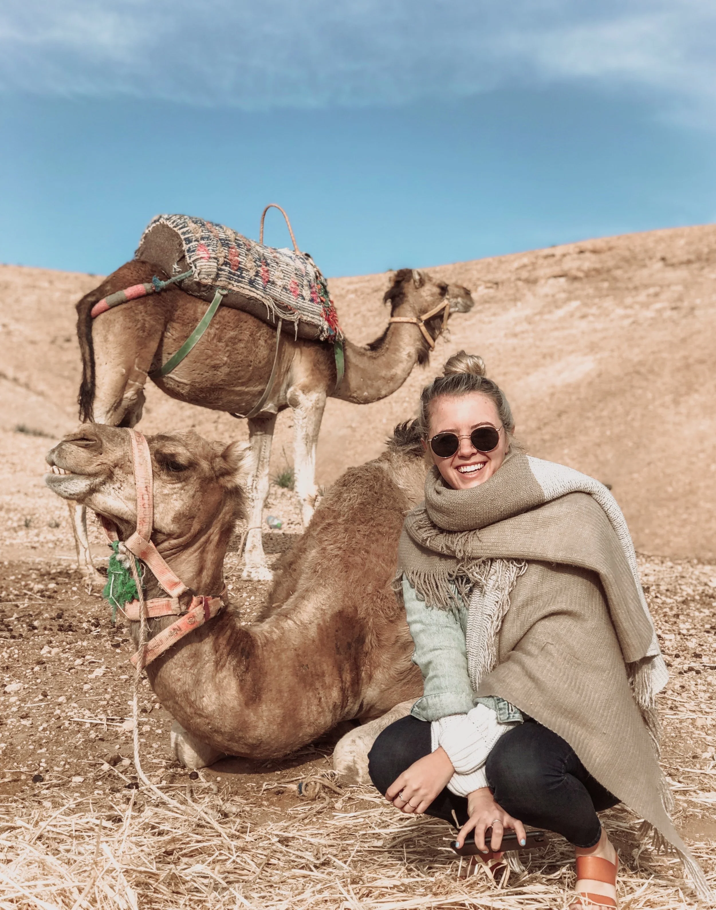 Woman crouching in desert terrain with two camels, one sitting and the other standing, under a blue sky.