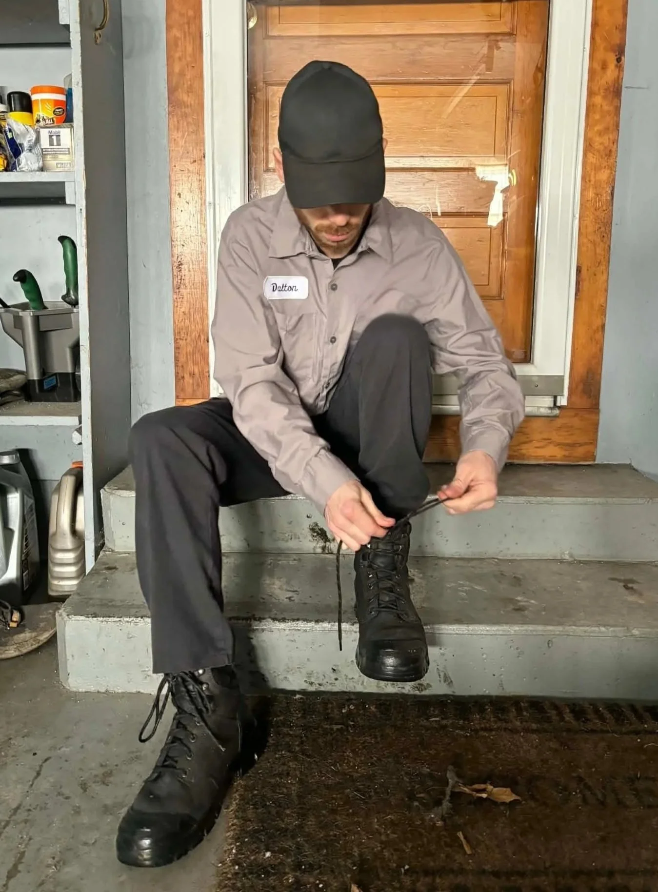 A man sitting on concrete steps, tying his black laced boots, in a garage or workshop area, wearing a gray uniform shirt with a name tag that says 'Dalton' and a black cap.