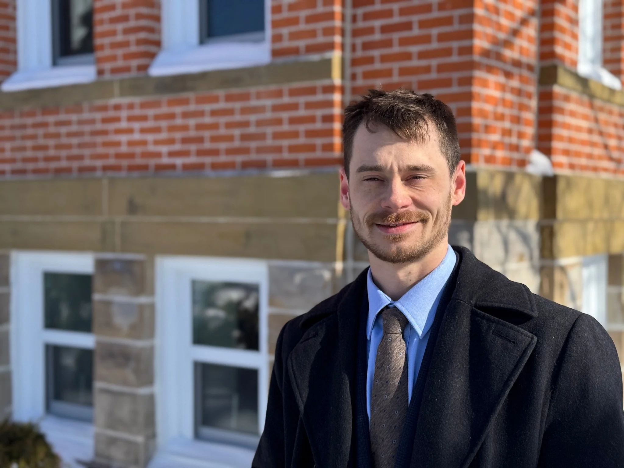 A young man in a suit and coat standing outside in front of a brick house with white-framed windows.