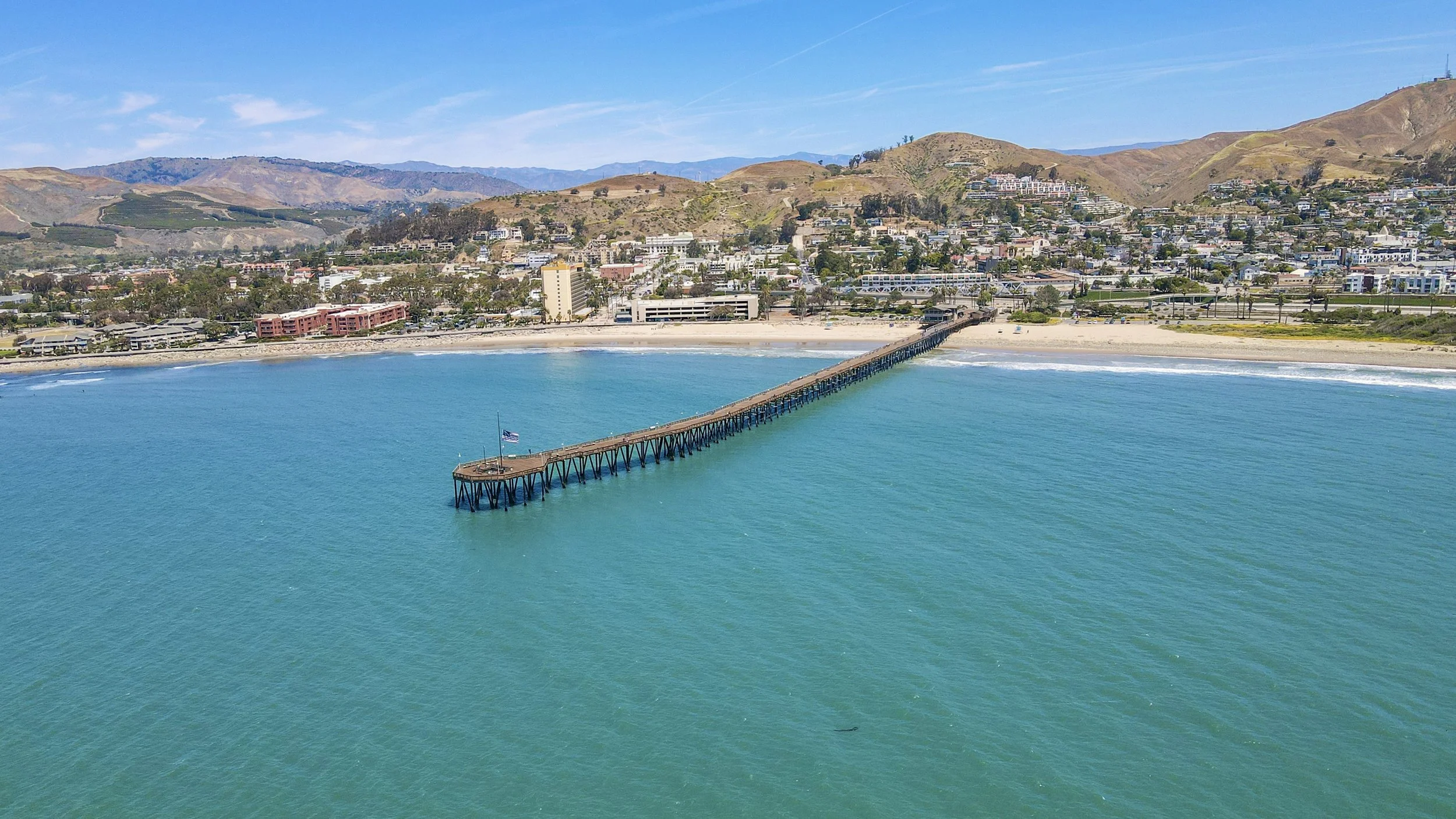 An aerial view of a pier extending into the ocean with a coastal town and hills in the background.