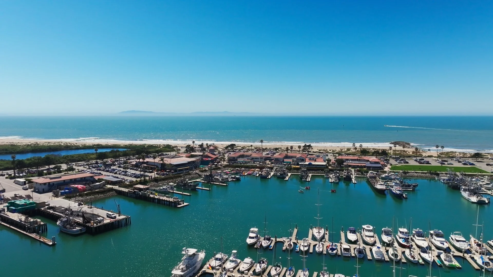 Aerial view of a marina with boats docked, a beach with sand, ocean waves, palm trees, and buildings along the shoreline, with a clear blue sky and a distant landmass on the horizon.