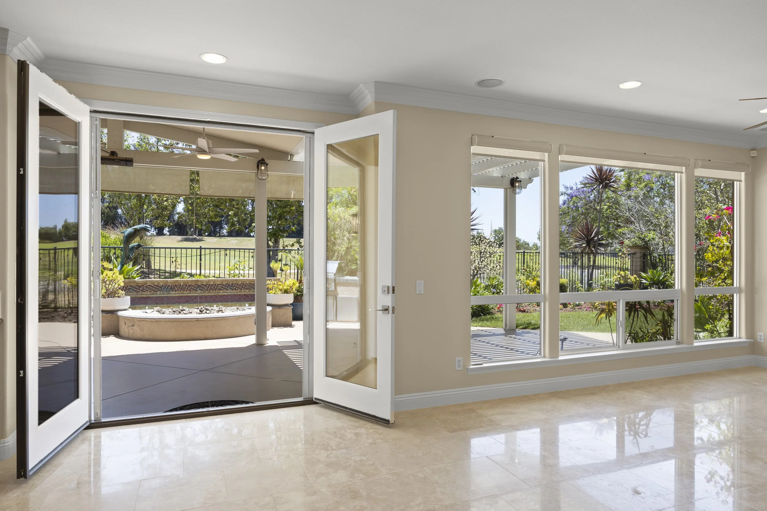 View of a bright indoor room with large windows and a sliding glass door opening to a backyard with a patio, garden, and outdoor decor.