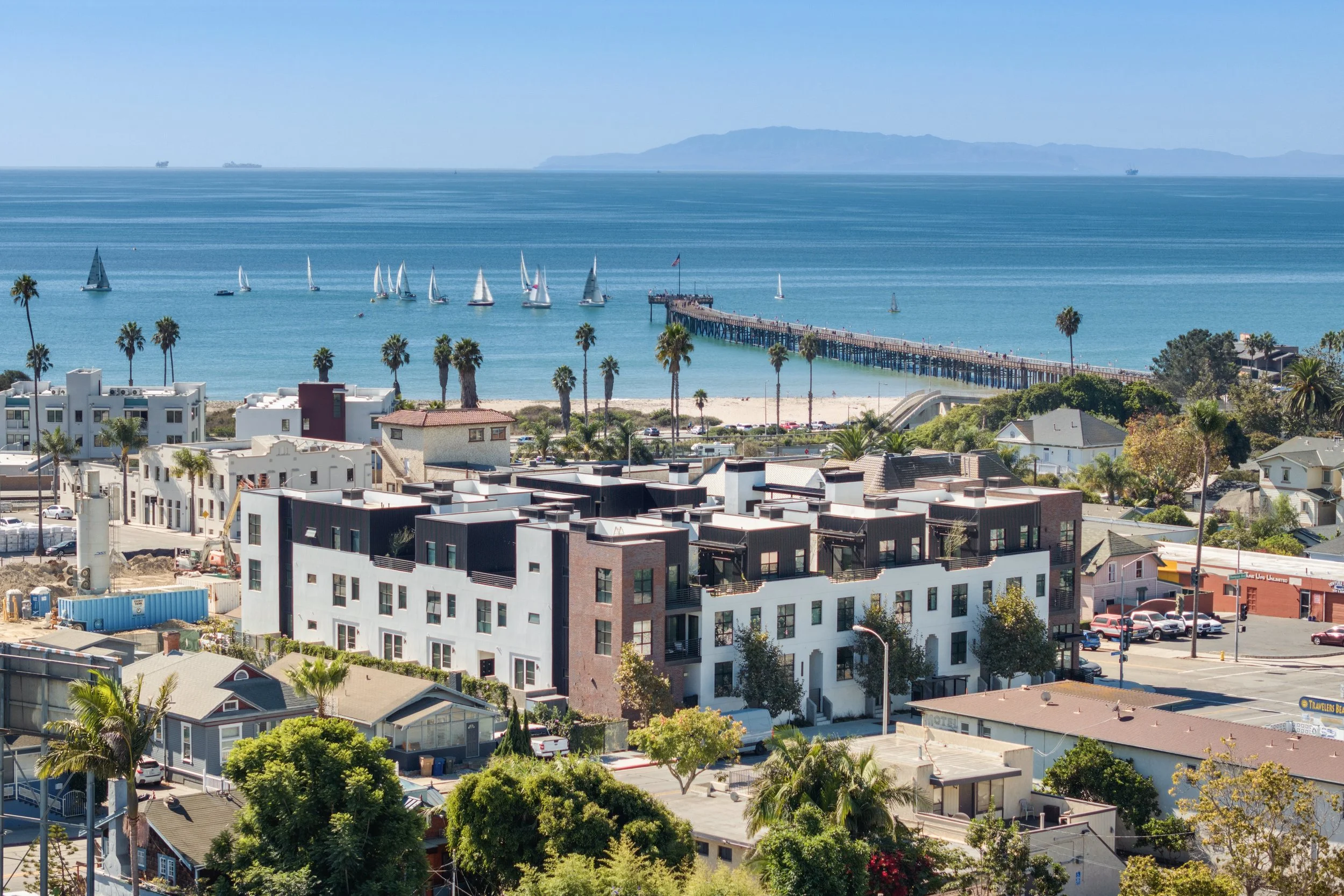 A coastal cityscape featuring modern white and dark-colored apartment buildings, lush green trees, and a view of the ocean with sailboats and a pier extending into the water, with a mountainous island in the background.