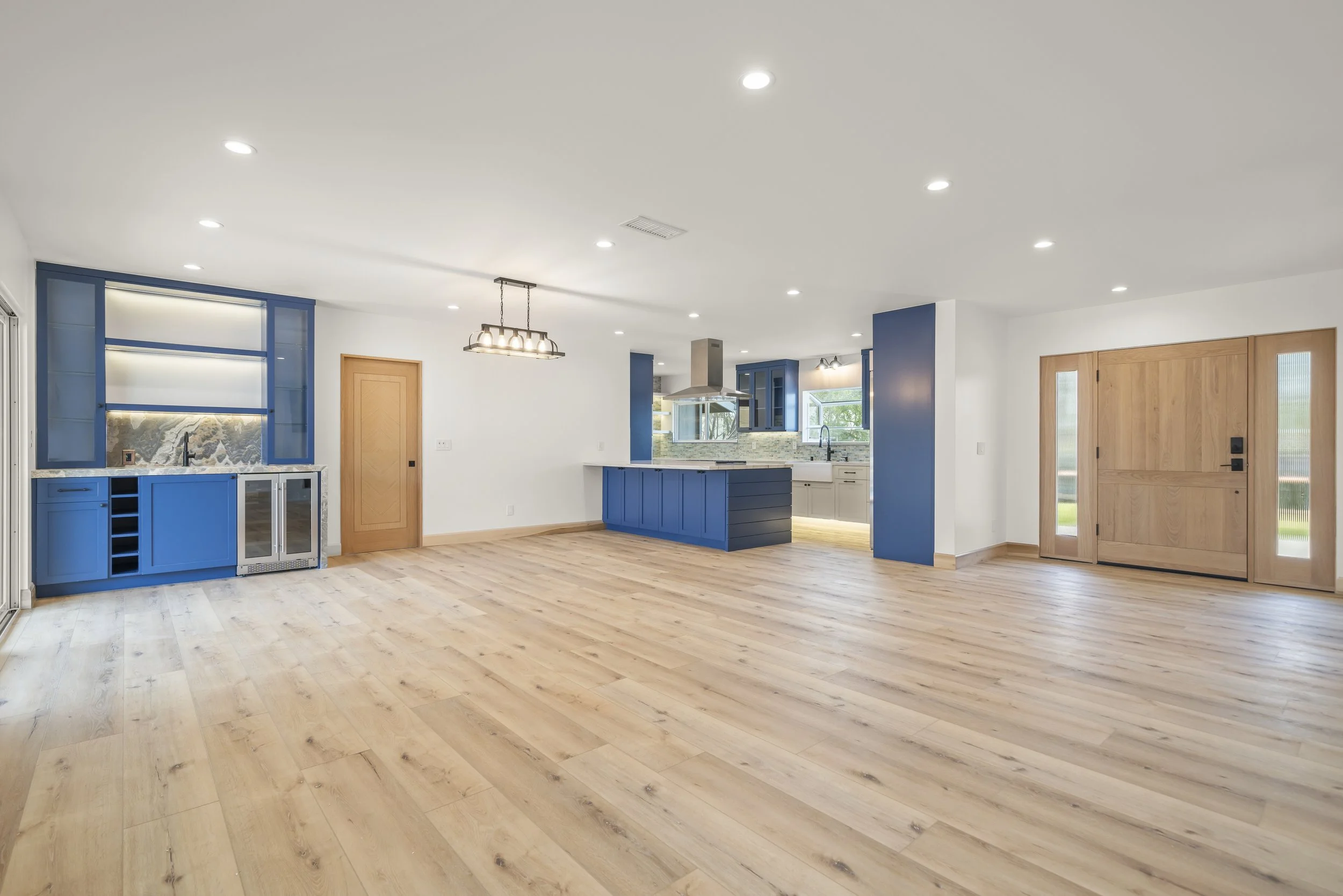 Empty open-concept living space and kitchen with light wood flooring, white walls, blue cabinetry, and natural light through windows.