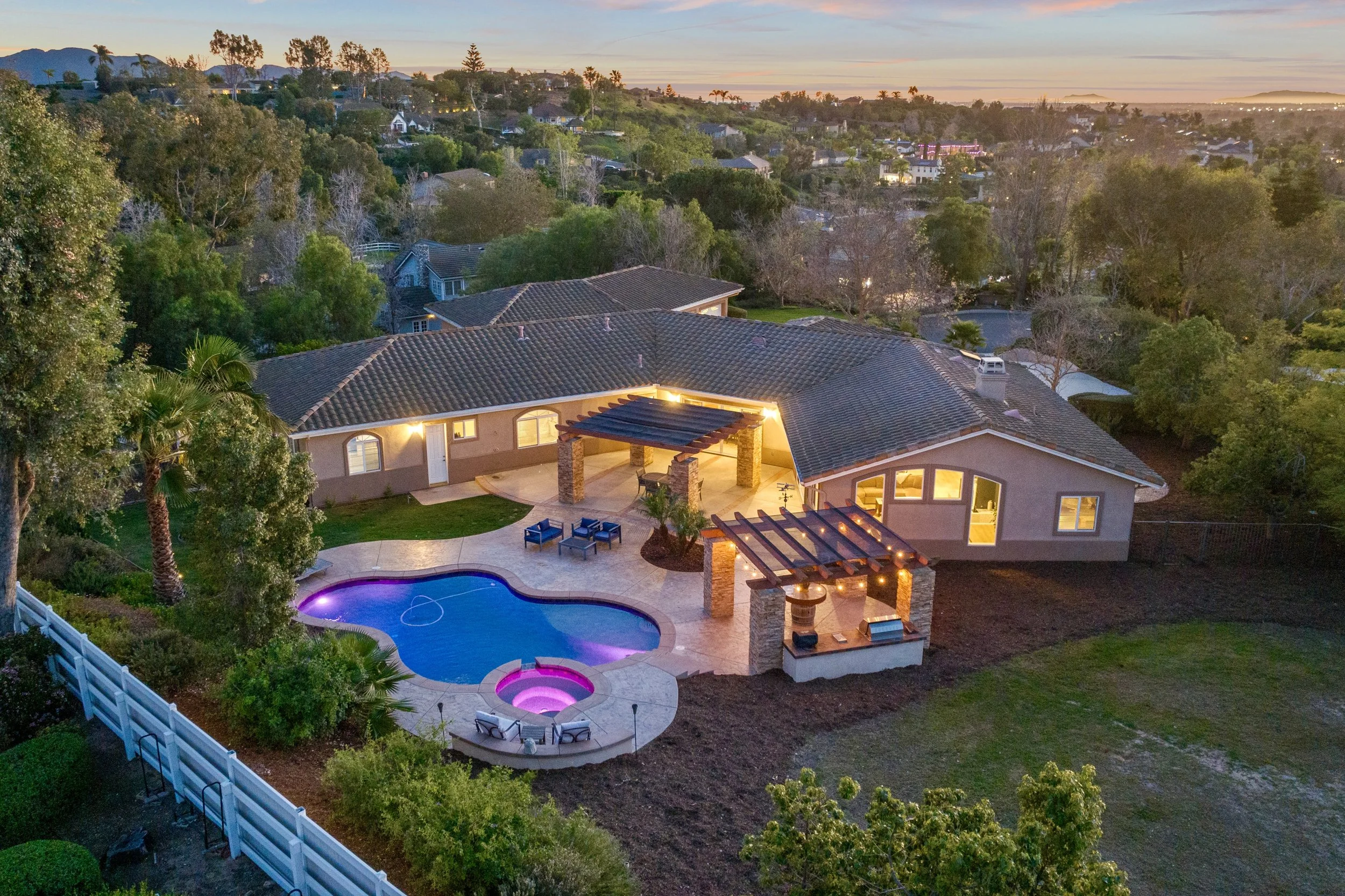 Aerial view of a backyard at sunset featuring a house with a tiled roof, outdoor seating area, illuminated swimming pool with purple lighting, hot tub with pink lighting, and surrounding trees and bushes.